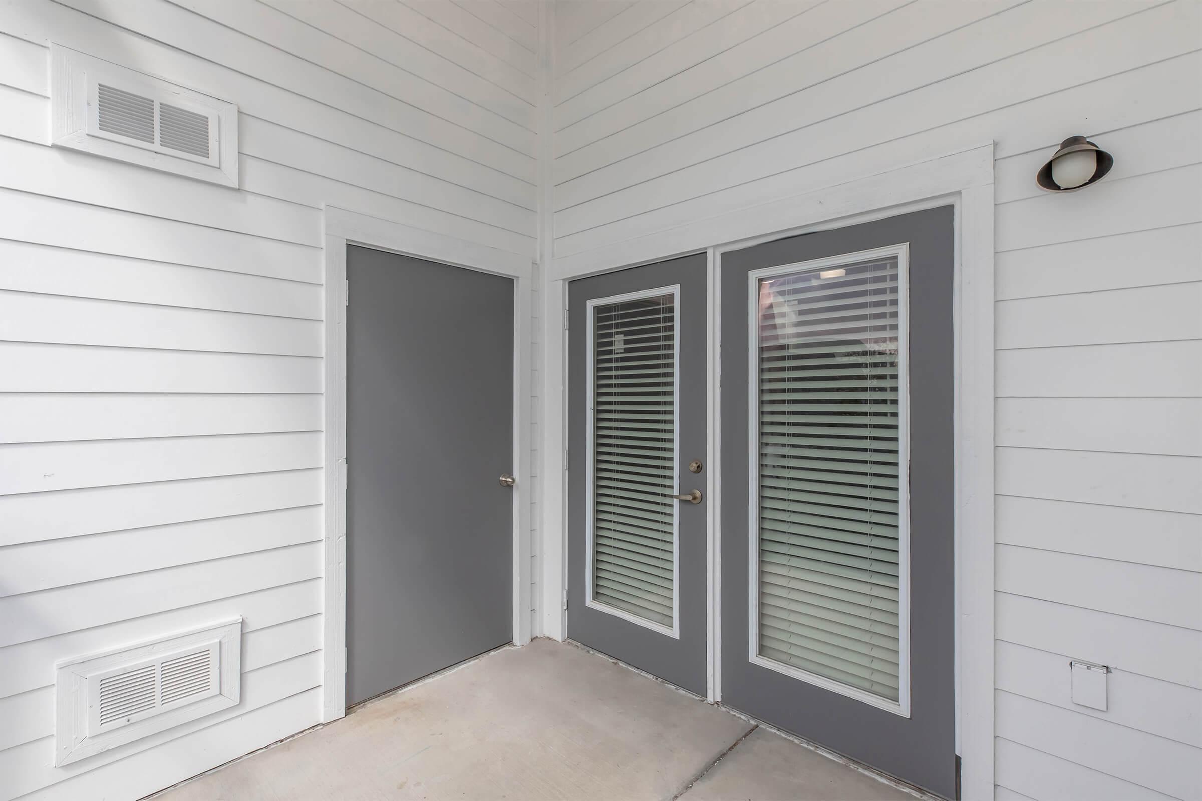 A corner entryway with two glass-paneled doors and a solid gray door. The walls are white with horizontal siding, and a light fixture is mounted on the wall. The concrete floor is visible, and there are vent openings near the ground. Natural light comes through the doors, illuminating the space.