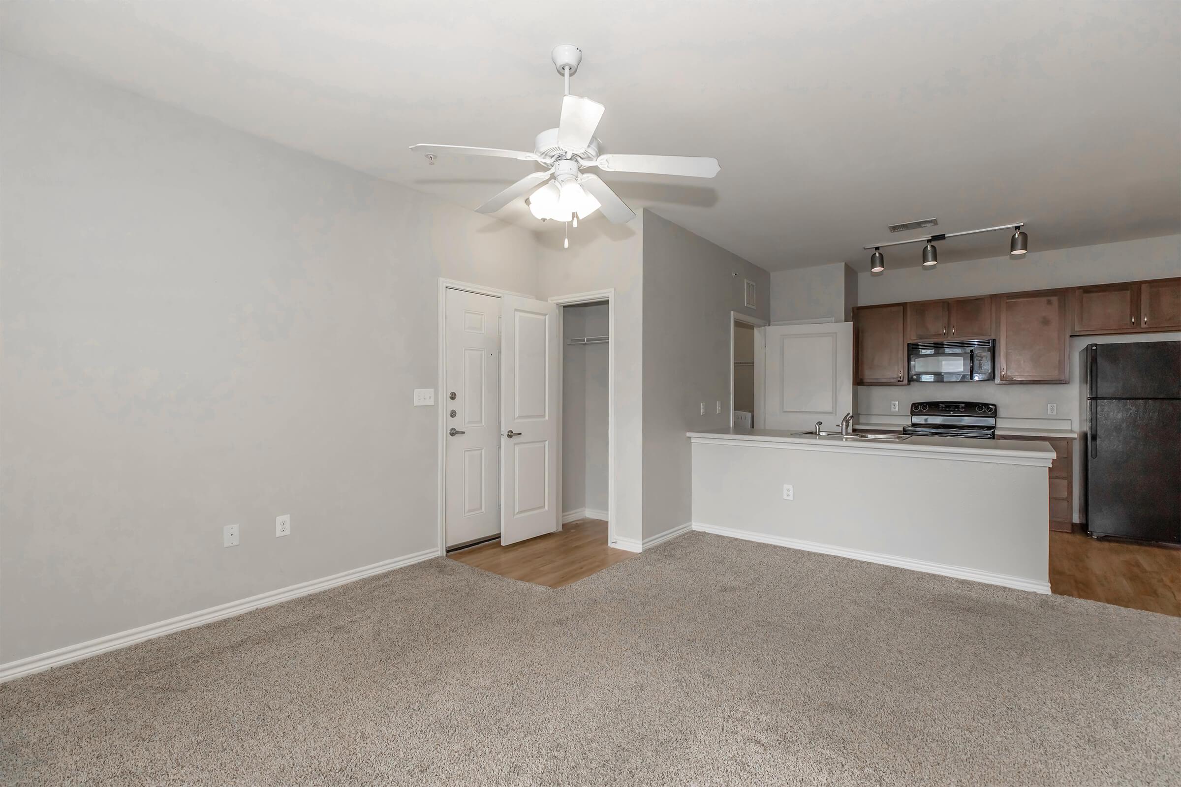 Spacious living area of an apartment featuring light-colored walls, a ceiling fan, and beige carpeting. The open layout includes a kitchen with wooden cabinets and a black refrigerator. An entrance door and closet can be seen to the left, creating a welcoming atmosphere. Bright and airy space perfect for relaxation and entertaining.