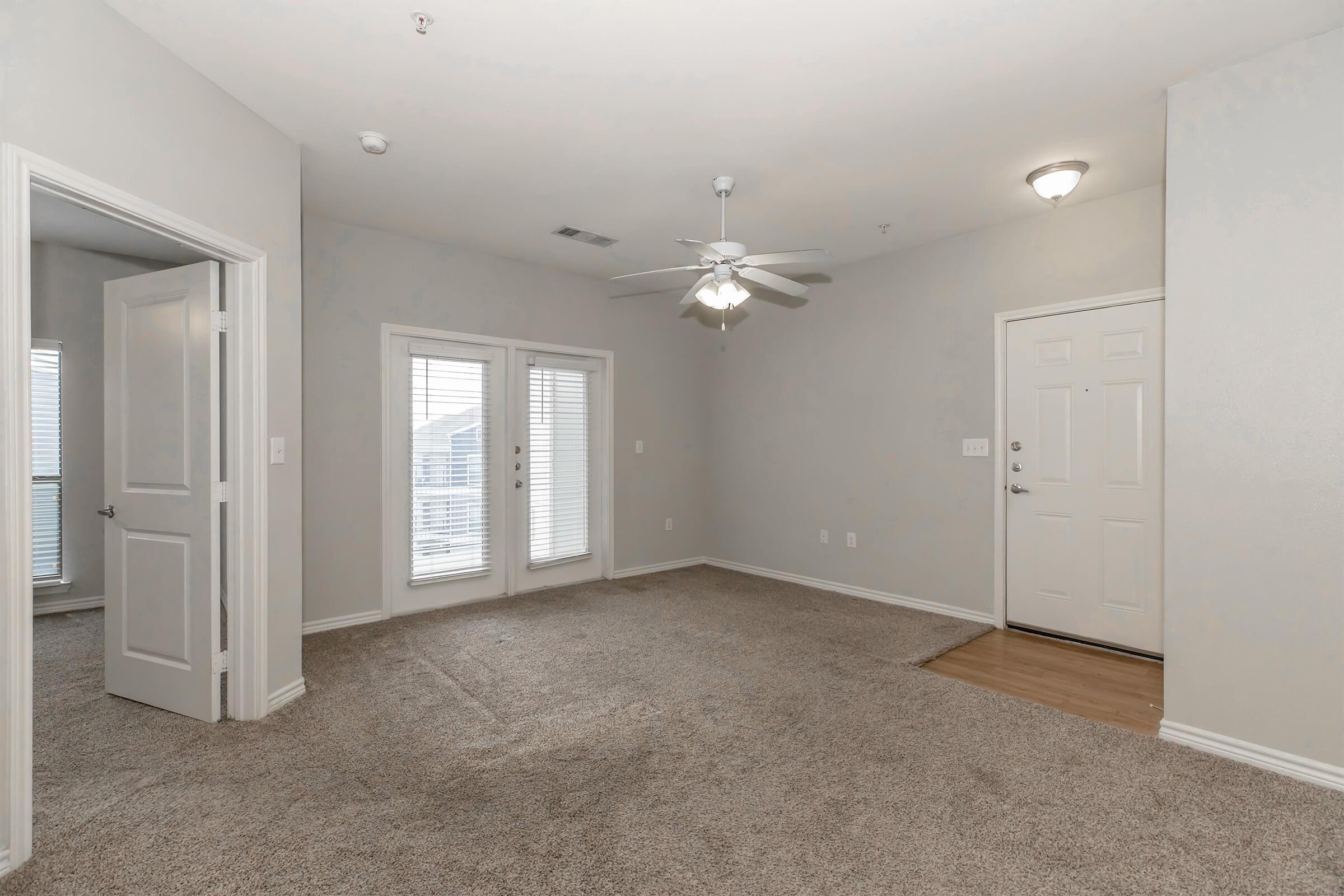 A spacious living room with neutral-colored walls and carpet. It features a ceiling fan, two windows with white shutters, and an entrance door. A second door leads to another room. The area is well-lit, giving a cozy atmosphere.
