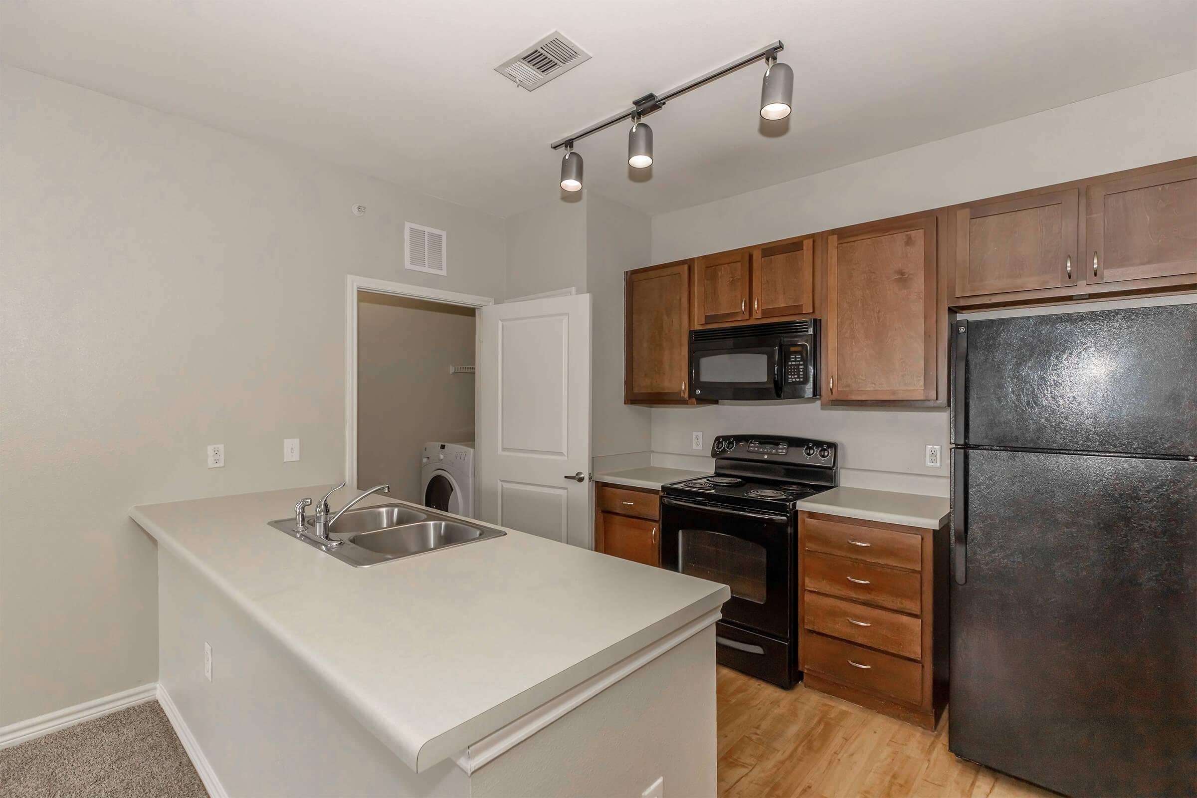 A modern kitchen featuring dark wood cabinets, a black refrigerator, and an electric stove. There is a double sink on the island, with pendant lighting above. The walls are painted a light color, and there is a door leading to a laundry area in the background. The flooring is laminate wood.