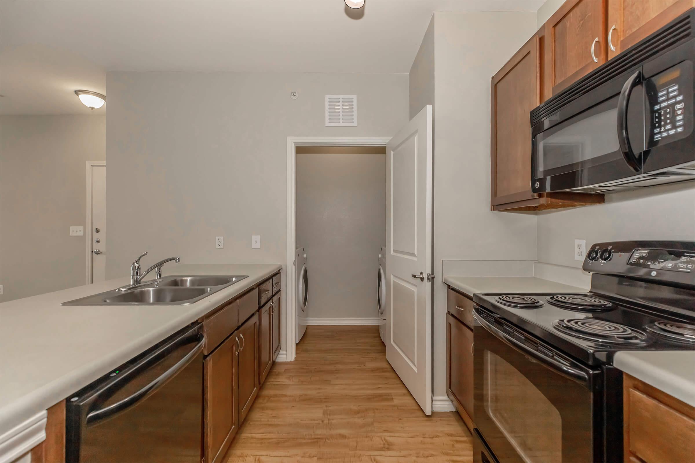 A modern kitchen featuring wooden cabinetry, a double sink, stainless steel appliances, and a black stove and microwave. The space has an open layout with light-colored walls and a doorway leading to a laundry area. A warm wood floor complements the design.