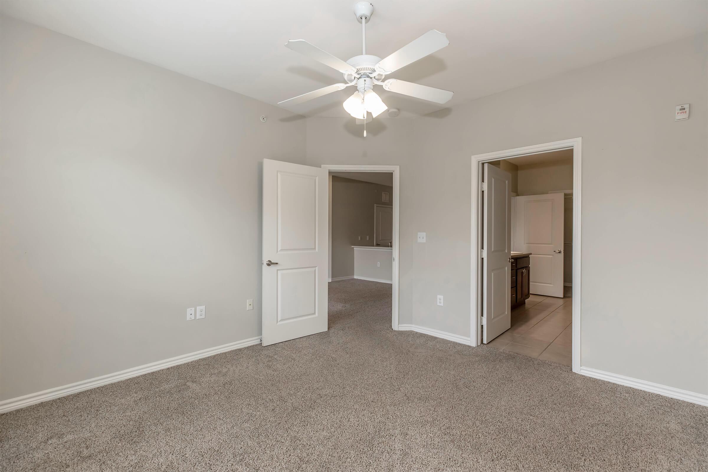A spacious, neutral-colored room featuring a ceiling fan and carpeted flooring. Two doorways lead to adjacent areas, one visible to a kitchen or hallway. The walls are painted light gray, creating a bright and airy atmosphere.