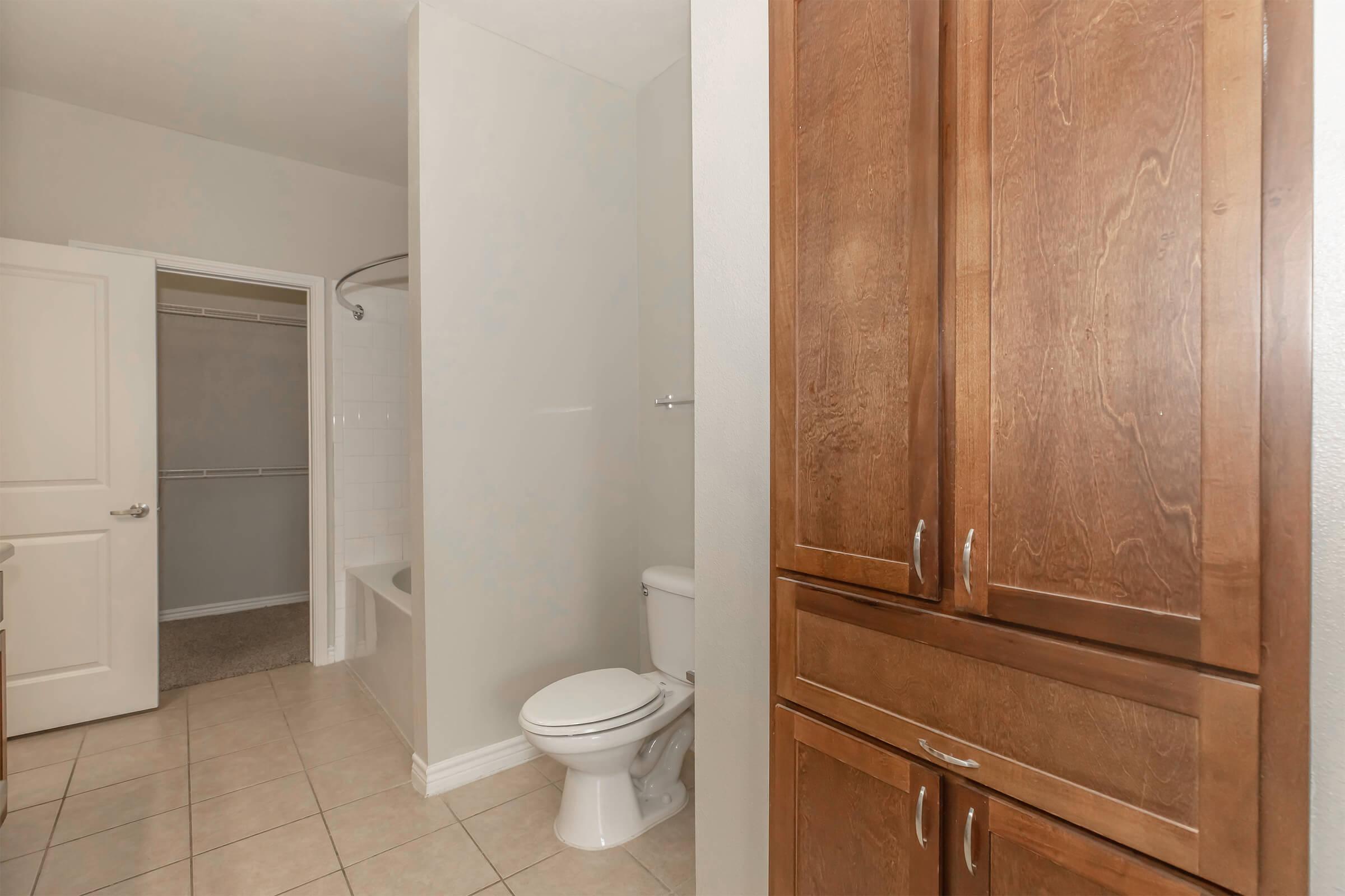 A view of a bathroom featuring a toilet, a tub, and a closet space. The space has light-colored walls and floor tiles, with wooden cabinets on the right side. The room appears clean and modern, with a simple layout.