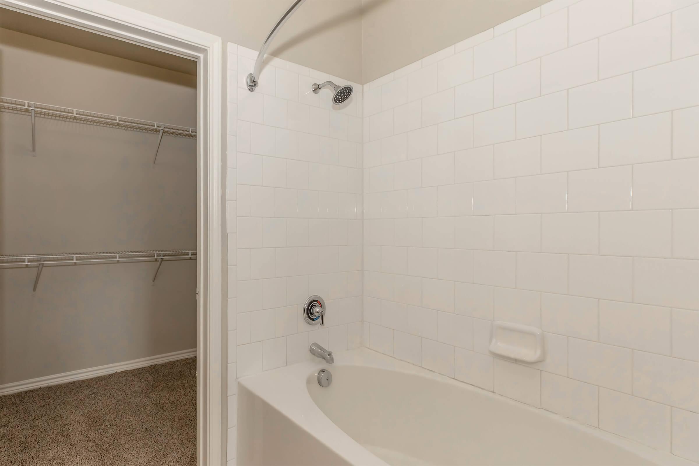 A clean, white-tiled bathroom featuring a bathtub with a shower fixture. The tub is positioned against a wall, and there is a sliding glass door opening to a closet with wire shelving. The floor is carpeted in a neutral color, and the walls are painted in a light shade.