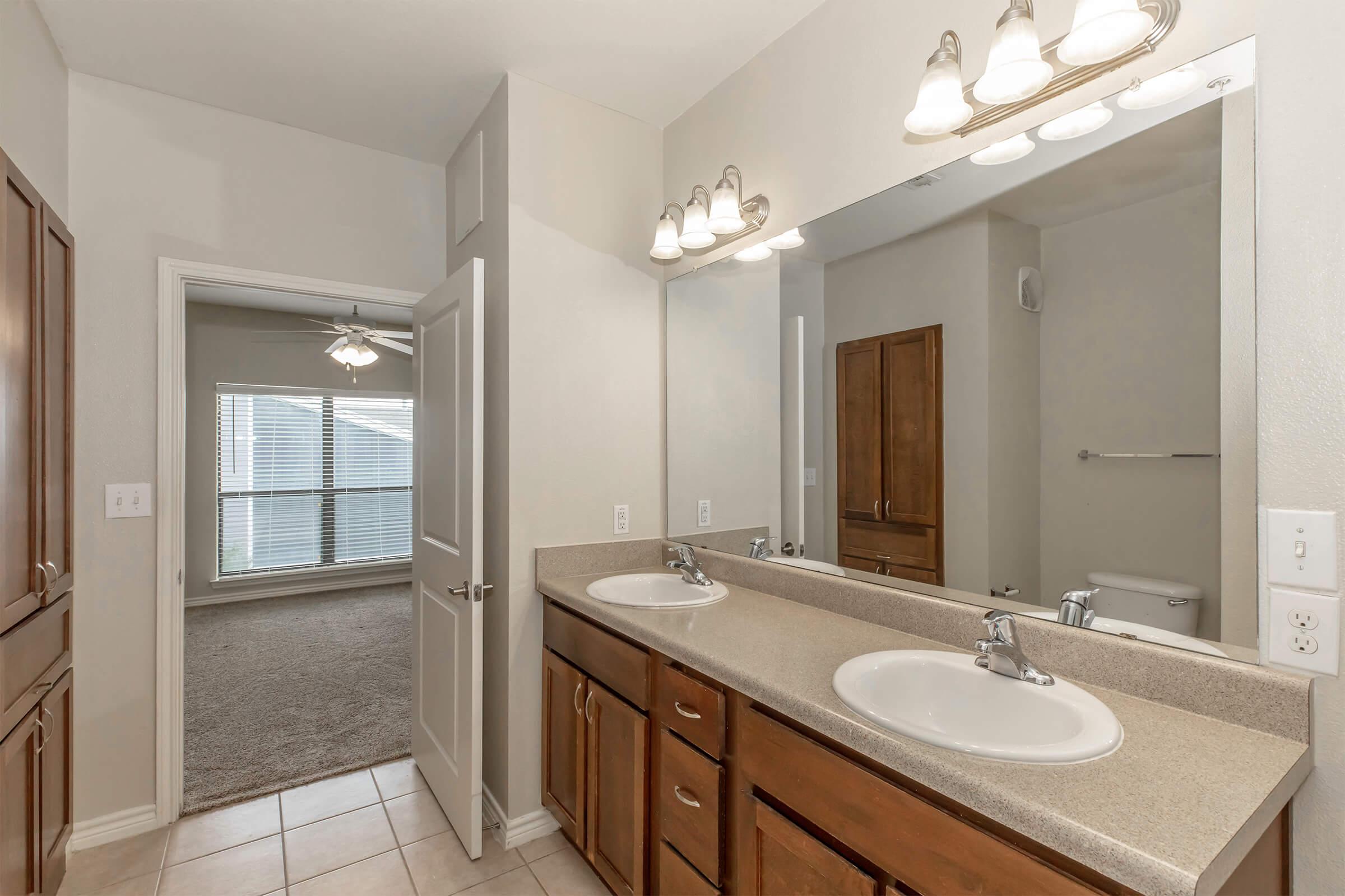 A well-lit bathroom featuring a double sink vanity with wooden cabinets, a large mirror with three light fixtures above. The room has light-colored walls and tile flooring, with a doorway leading to a bright, carpeted area visible in the background.