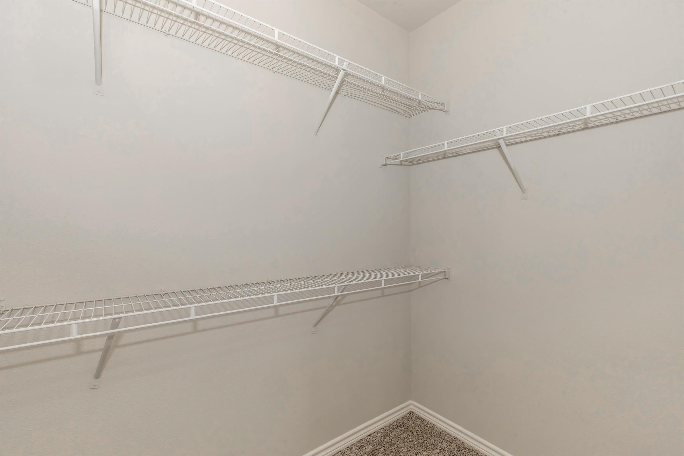 Empty closet with white wire shelving on the walls, showcasing a light-colored interior and carpeted floor. The shelves are positioned at different heights, providing a spacious and organized storage area.