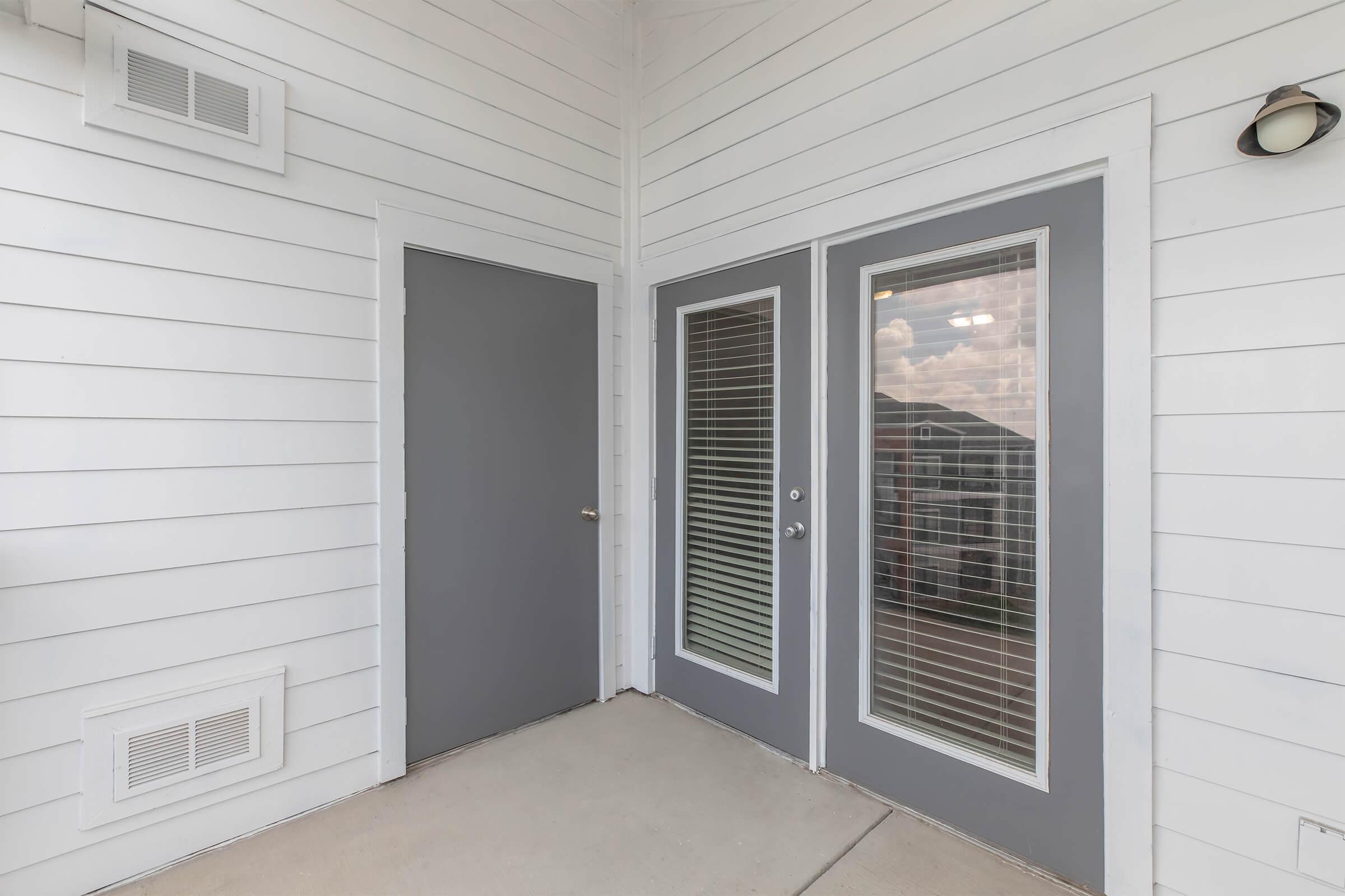 A modern entryway featuring a grey door to the left and double glass doors on the right, both with horizontal blinds. The exterior walls are white with horizontal siding, and there is a light fixture on the right side of the entrance. The floor is concrete, creating a clean, minimalist look.