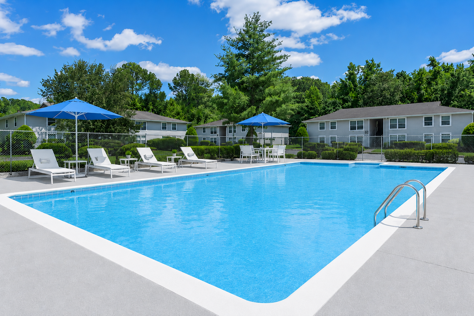 A clear blue swimming pool surrounded by lounge chairs and blue umbrellas. In the background, there are several single-story buildings and lush green trees, under a bright blue sky with fluffy white clouds. The pool area is well-maintained, featuring a gradual entry with a ladder.