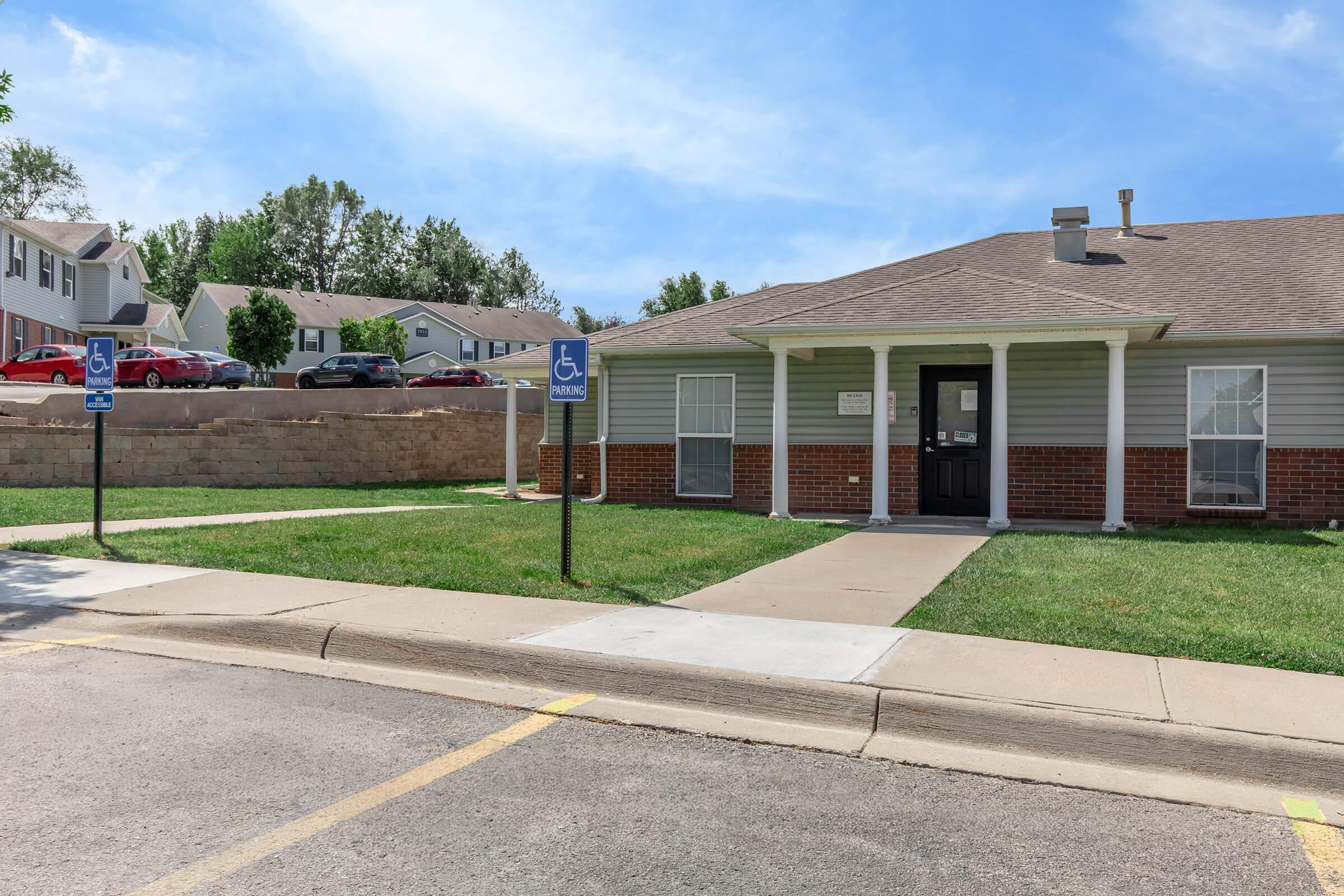 A single-story building with a sloped roof and brick accents, surrounded by grass and a concrete pathway. There are designated parking spots for individuals with disabilities in the foreground. In the background, several residential buildings are visible against a clear blue sky.
