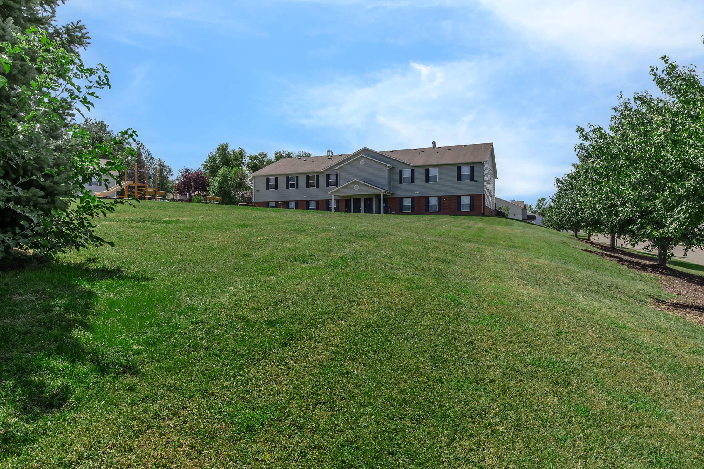 A spacious green lawn sloping gently toward a two-story building with multiple windows and a covered entrance. Surrounding the property are trees, creating a peaceful and inviting outdoor space under a clear blue sky.