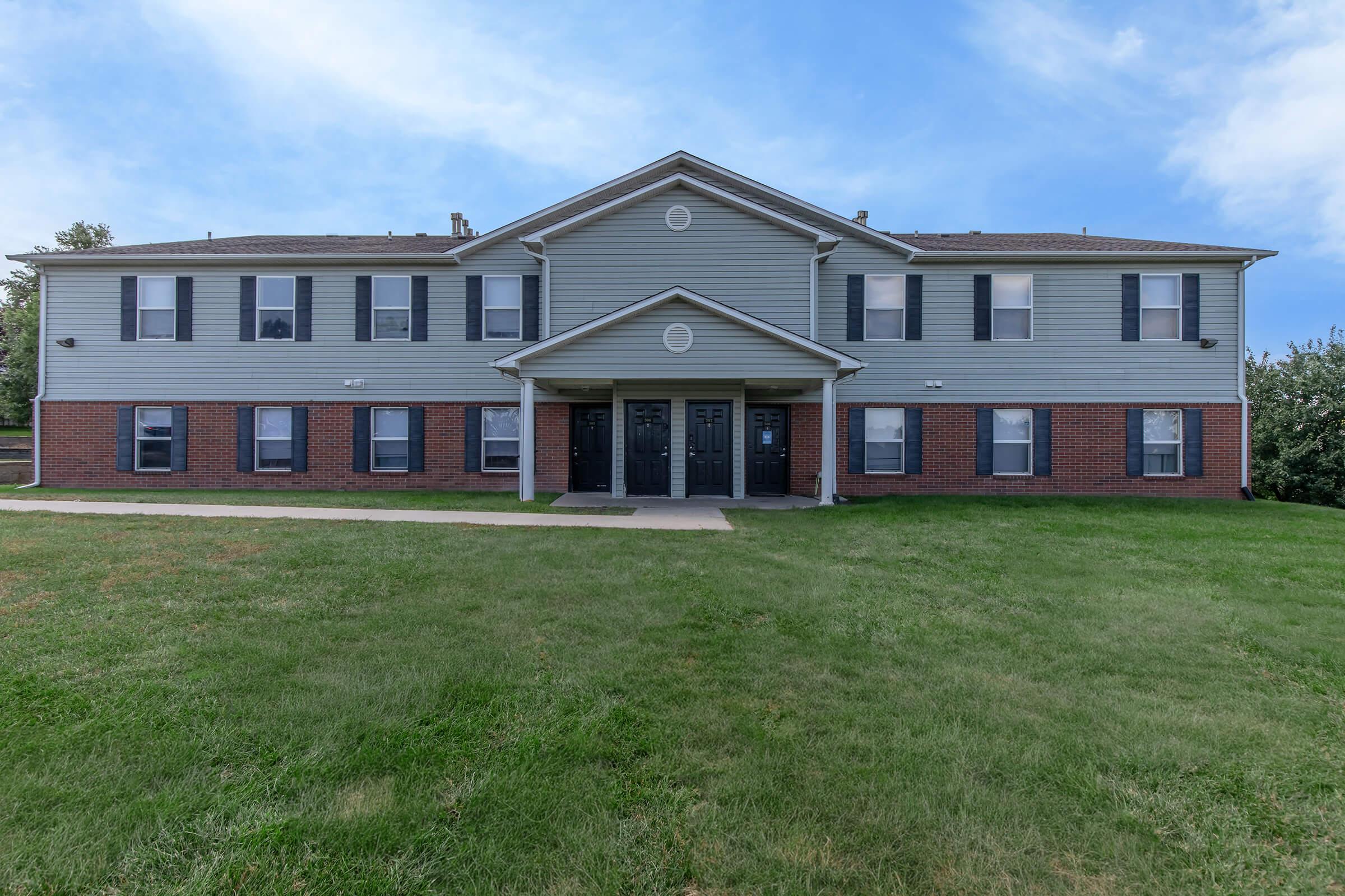 A two-story residential building with a light green exterior and brick accents. The building features several windows with black shutters and three entrance doors on the front. It is surrounded by a well-maintained grassy lawn under a partly cloudy sky.