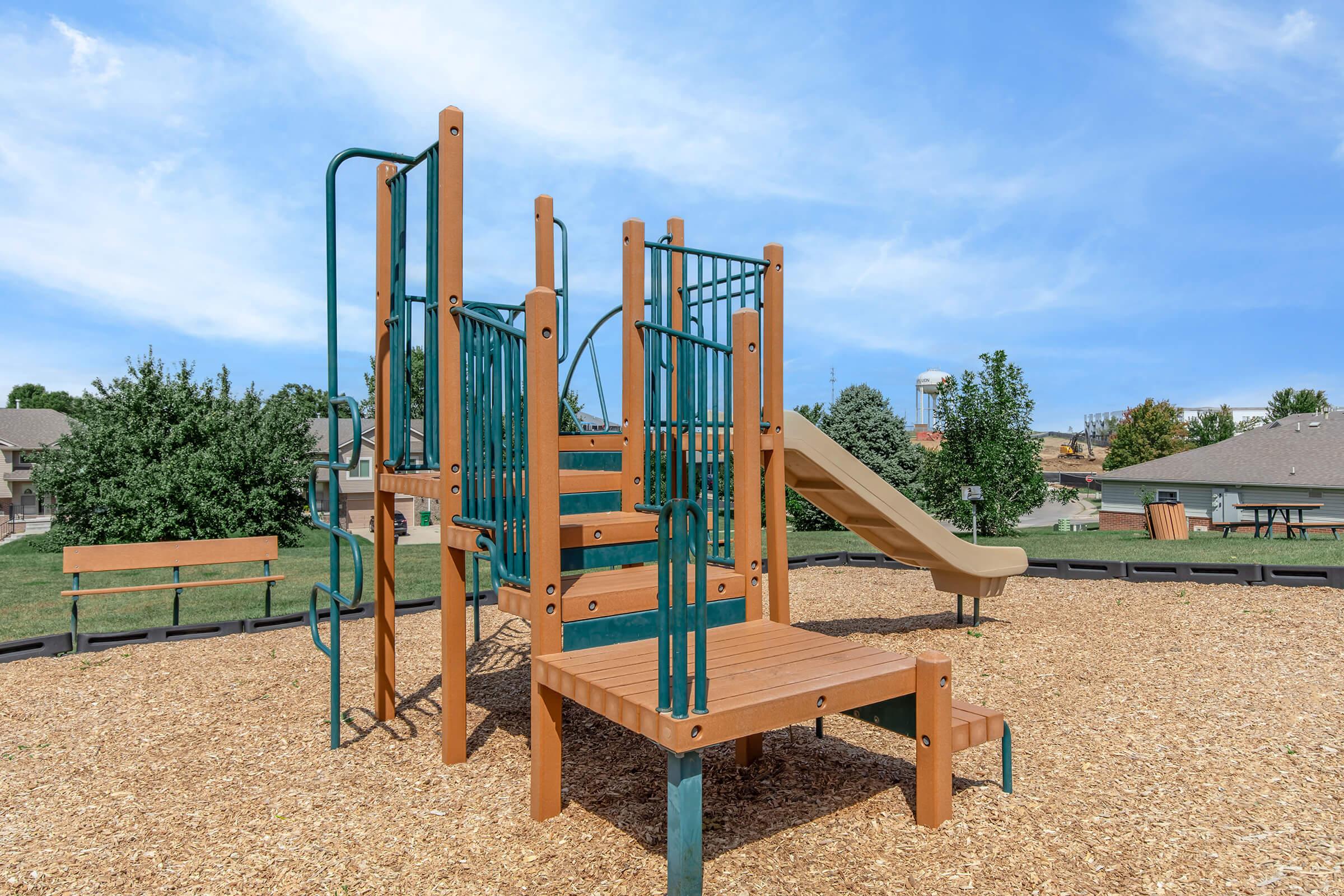 Playground structure featuring a climbing frame with stairs and a slide, surrounded by wood chip flooring. The scene includes grassy areas and trees in the background under a clear blue sky, offering a welcoming environment for children to play and explore.