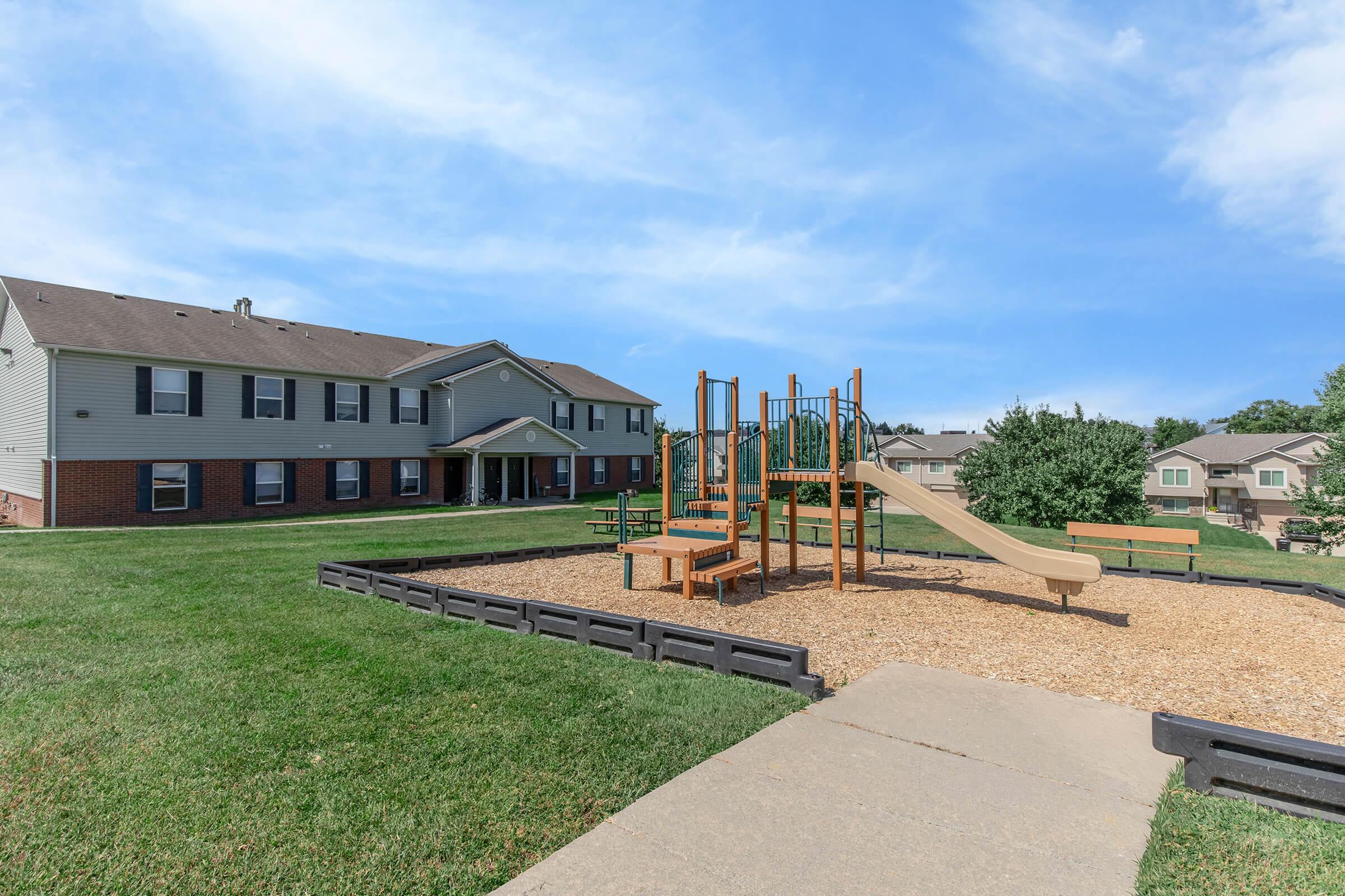 A playground with a slide and climbing structure sits on a grassy area, surrounded by gravel. Nearby, there are benches and a concrete path leading towards a multi-story residential building in the background under a clear blue sky.