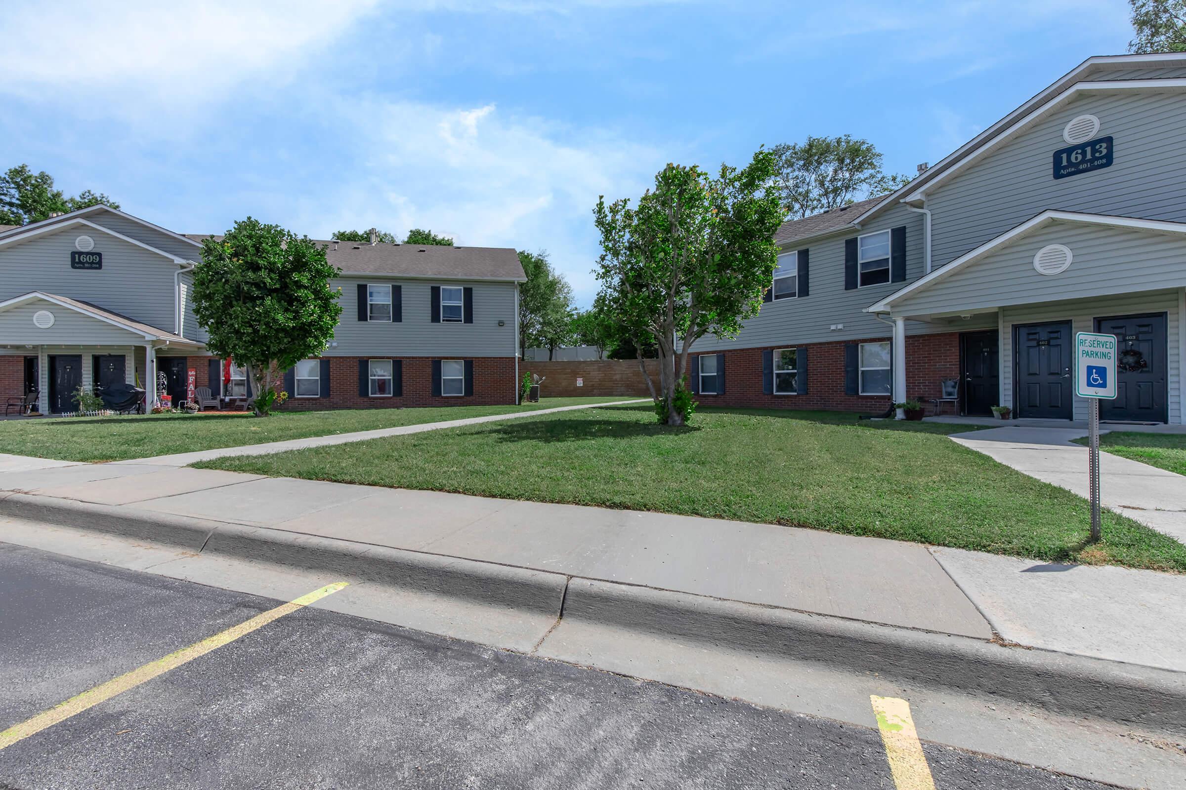 Two-story residential buildings with light gray siding and red brick accents. The foreground features a grassy area with small trees. A parking lot runs alongside the buildings, and a sign indicates accessible parking. The setting is bright and sunny, with a clear sky in the background.