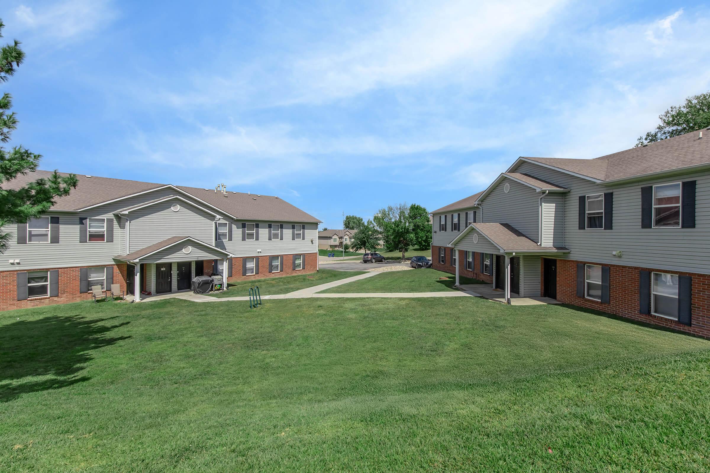 A serene view of a residential complex featuring two-story buildings with beige siding and brick accents. The buildings are surrounded by well-maintained green lawns and pathways, under a clear blue sky. Trees are visible in the background, adding to the tranquil atmosphere.