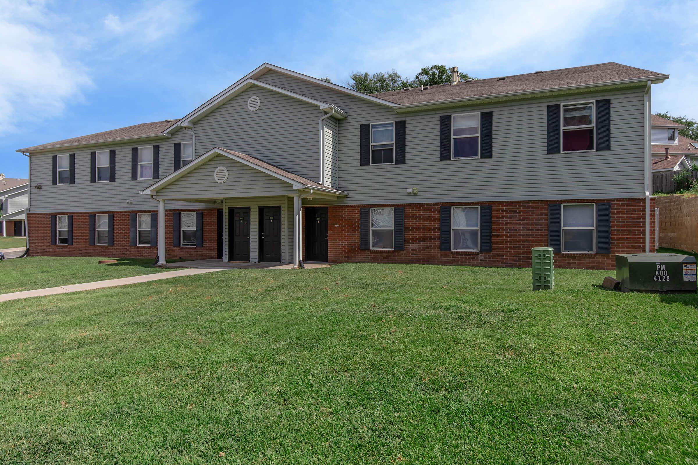 A two-story residential building featuring a brick and siding exterior. The facade includes a central entrance with two doors, surrounded by windows. The building is set on a grassy lawn with a clear blue sky above, creating a pleasant environment.