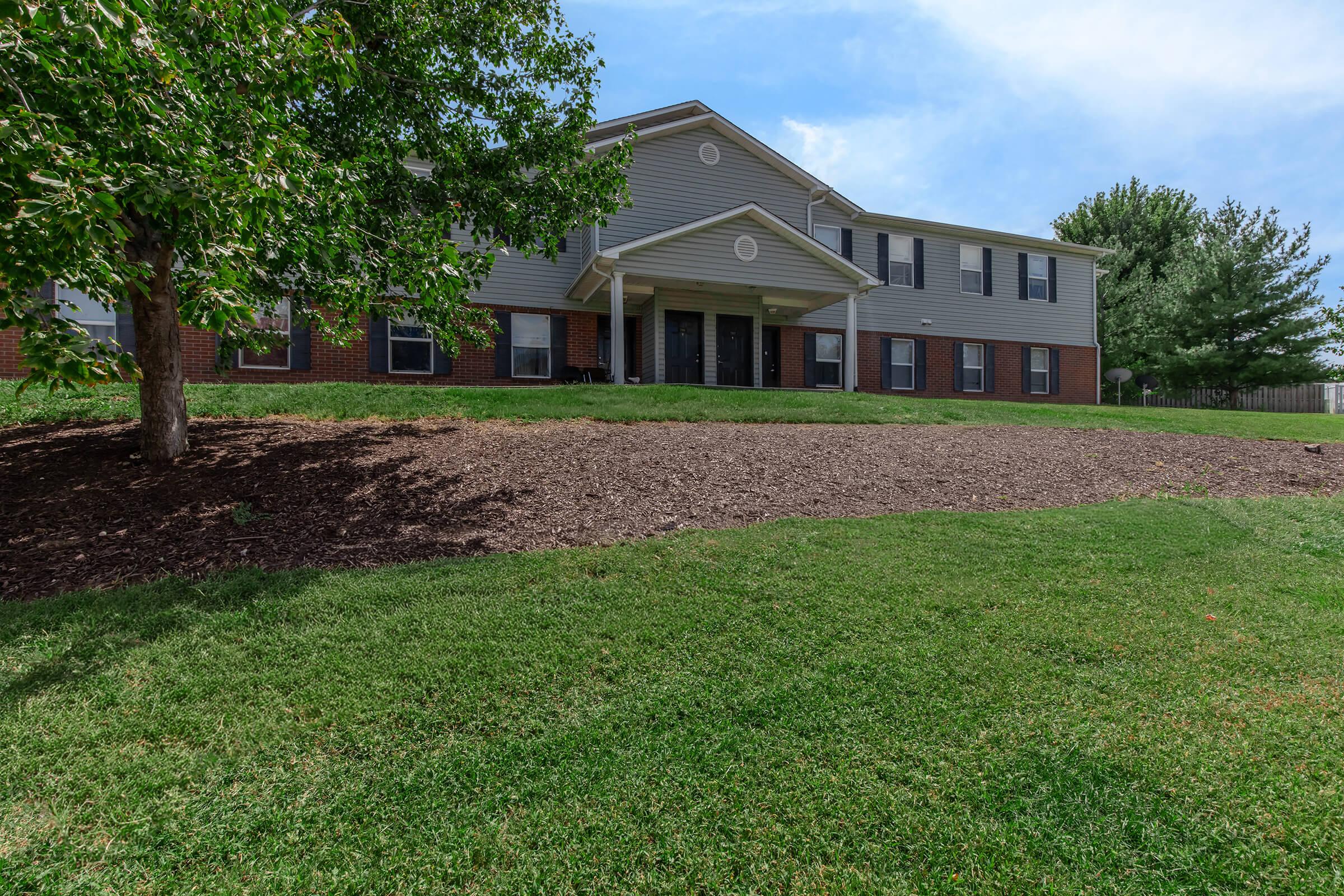 A two-story residential building with a light gray exterior and black doors, set against a backdrop of green grass and a landscaped area featuring mulch and a tree. The building has multiple windows and a manicured lawn. Clear blue sky is visible above.
