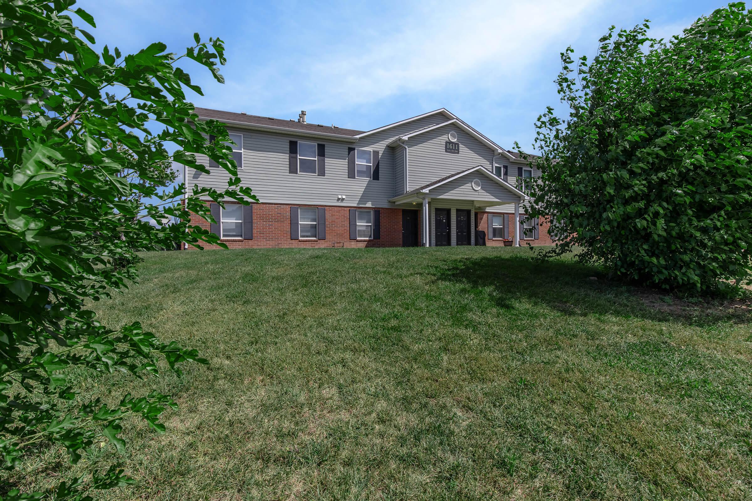 A two-story apartment building with a brick and gray siding exterior, surrounded by a grassy lawn and small trees or shrubs in the foreground. The sky is clear with a few clouds. The building features multiple windows and a central entrance.