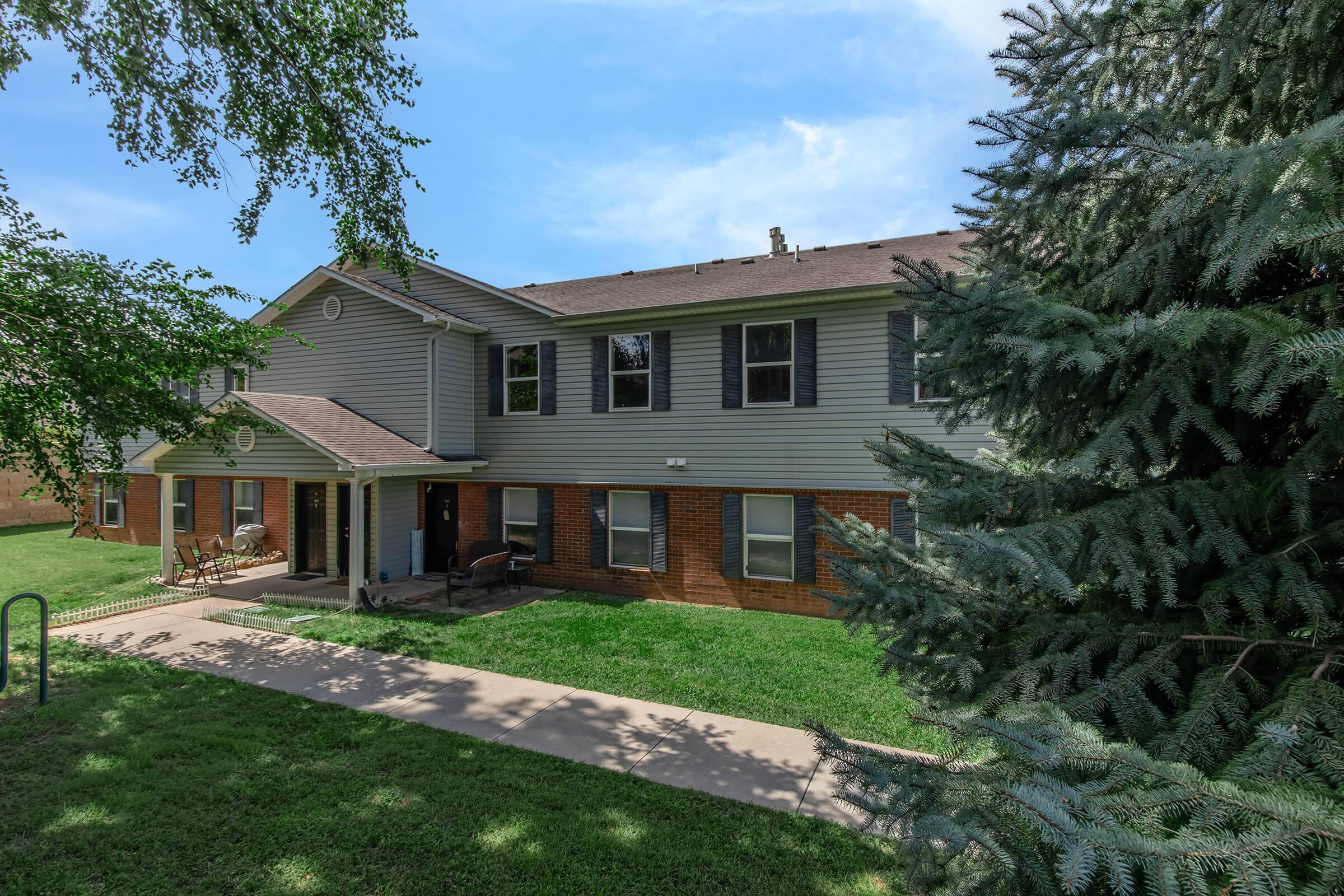 Two-story residential building with a gray exterior and brick accents, surrounded by greenery. There is a walkway leading to the front entrance, and some chairs are visible on the porch. A tree partially frames the scene in the foreground under a clear blue sky.