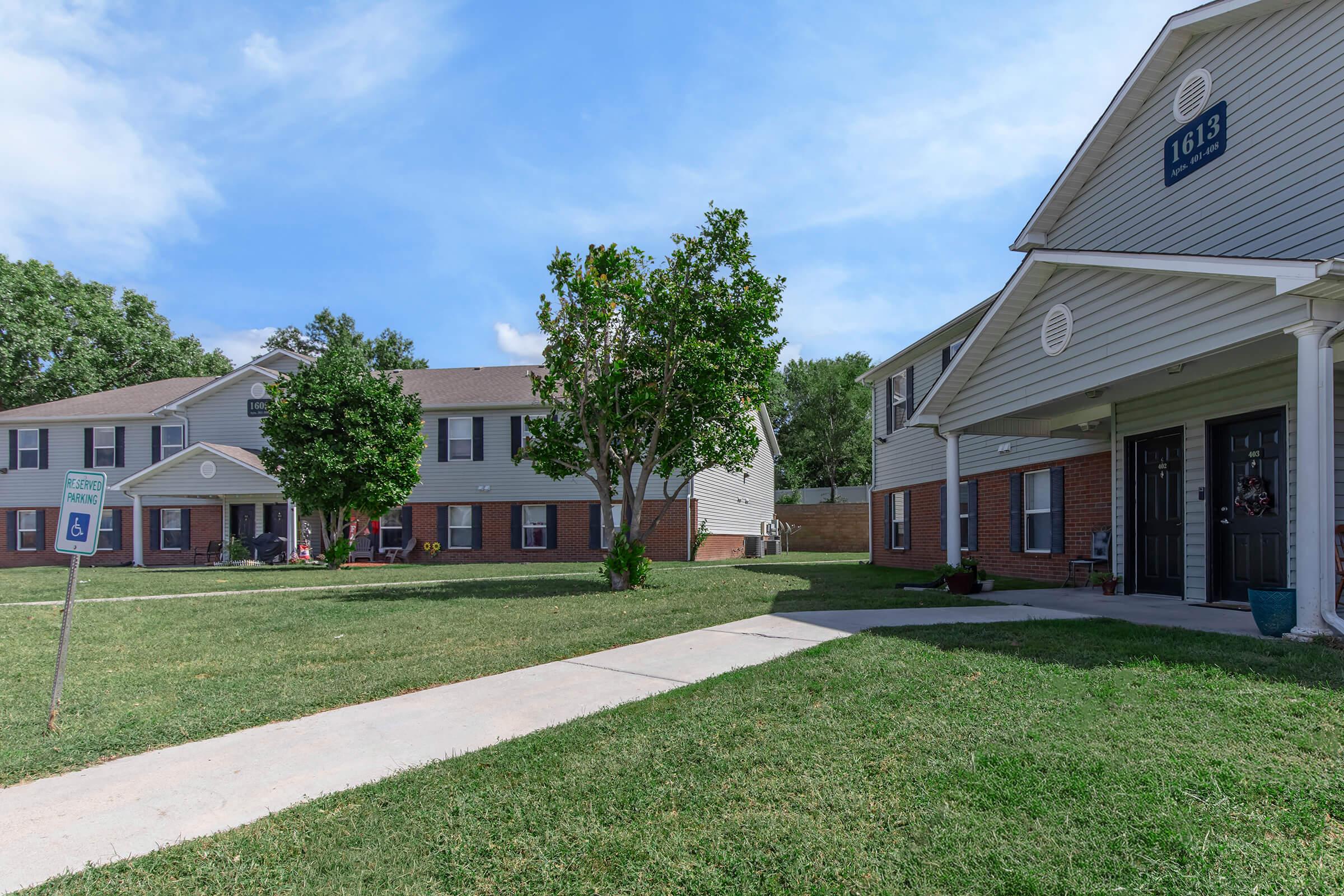 A residential community featuring two-story apartment buildings with gray siding, green grassy areas, and small trees. A sidewalk leads through the property, and a sign indicating accessible parking is visible in the foreground. The sky is clear with a few clouds.