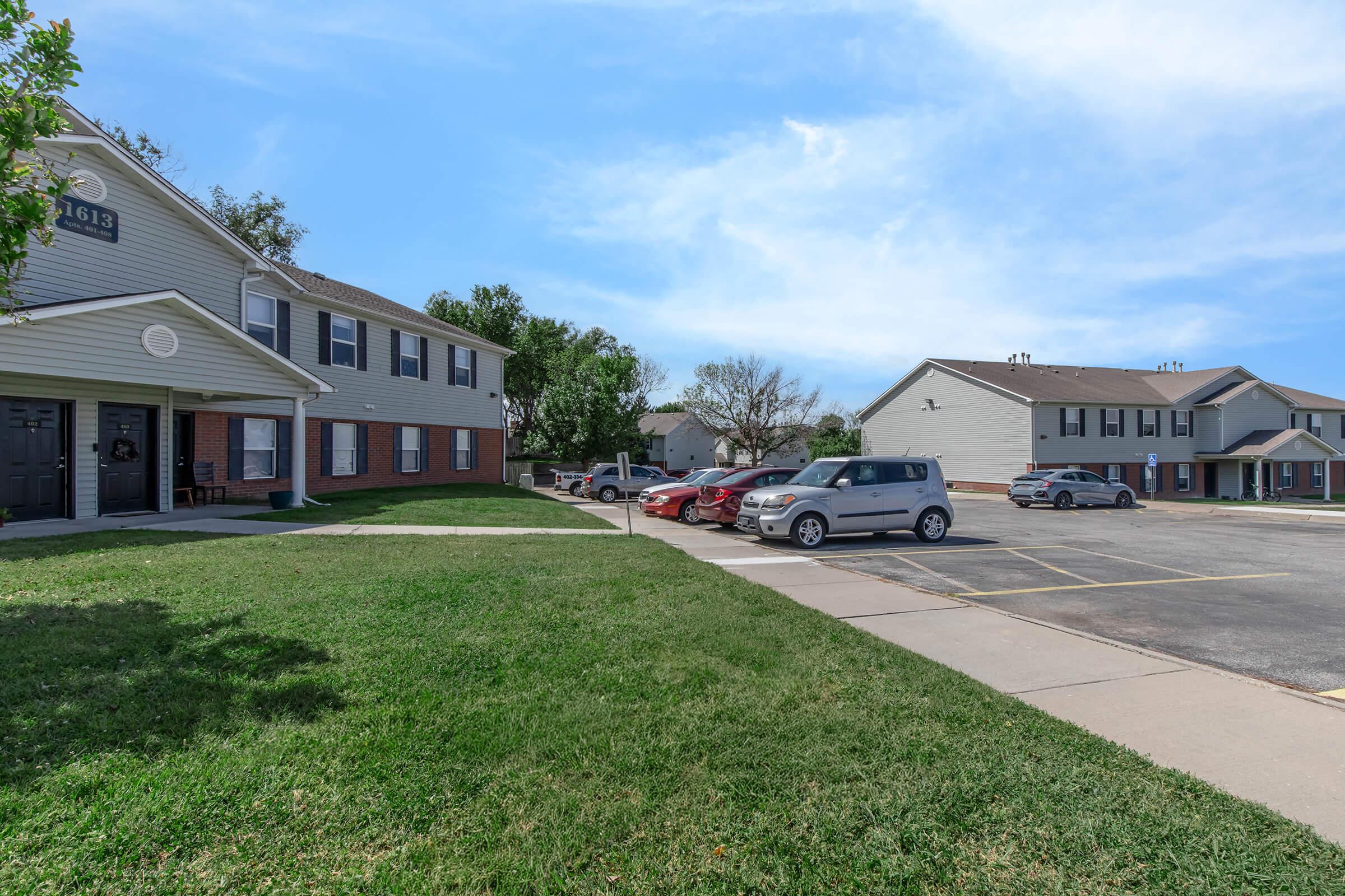 A suburban apartment complex featuring two buildings. The foreground shows a grassy area with a parking lot where several cars are parked. One building has a porch area, and the sky is clear with few clouds. The scene conveys a peaceful residential atmosphere.