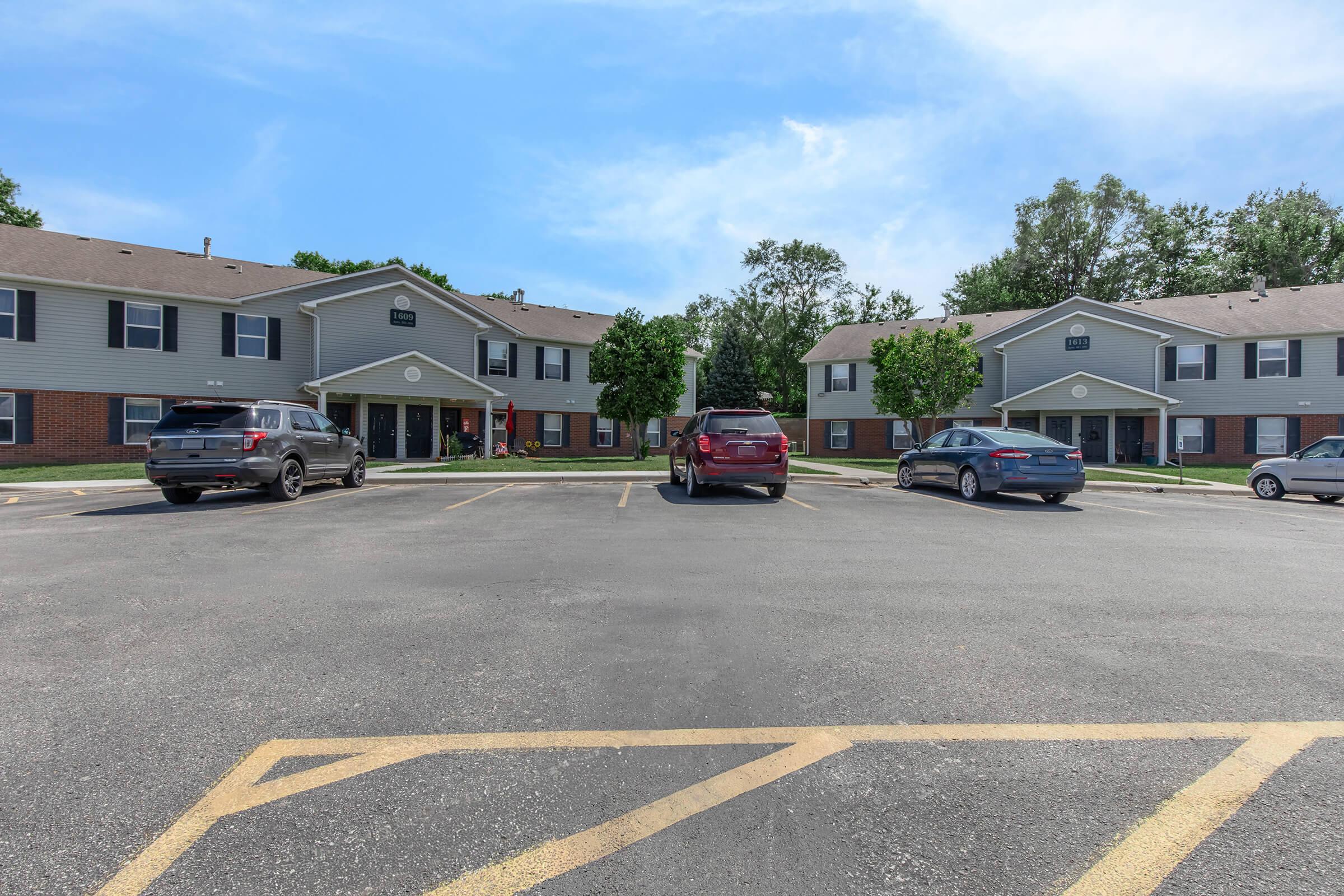 A residential parking lot with several parked cars in front of two-story apartment buildings. The buildings have gray siding, with some trees and greenery nearby. The sky is clear and blue, indicating a sunny day.