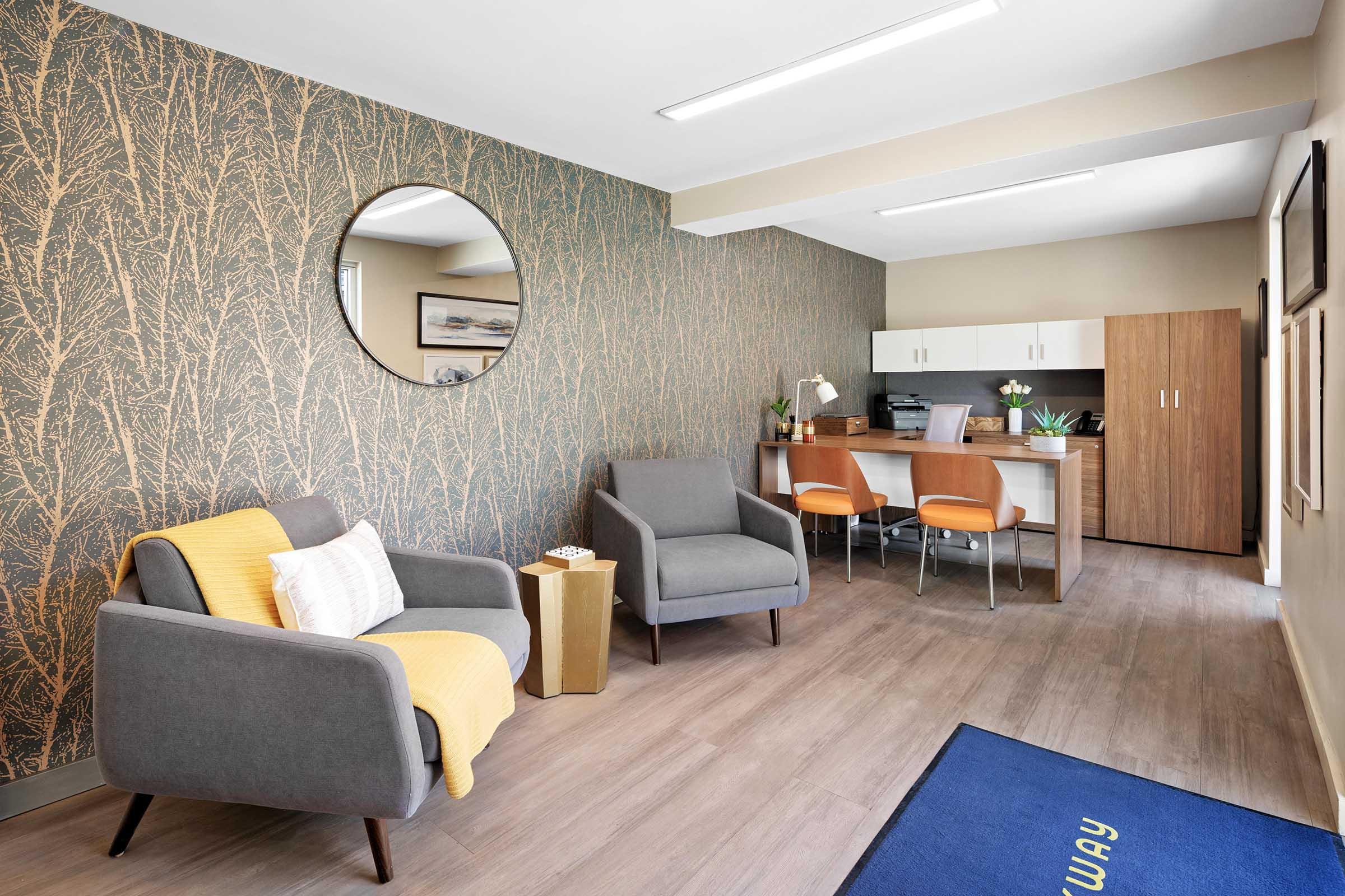 Modern office space featuring a cozy seating area with gray armchairs and a yellow throw blanket. A round mirror adorns the wall, which has a nature-inspired pattern. In the background, a desk area with a computer and office supplies is visible, along with wooden cabinetry and a blue welcome mat.