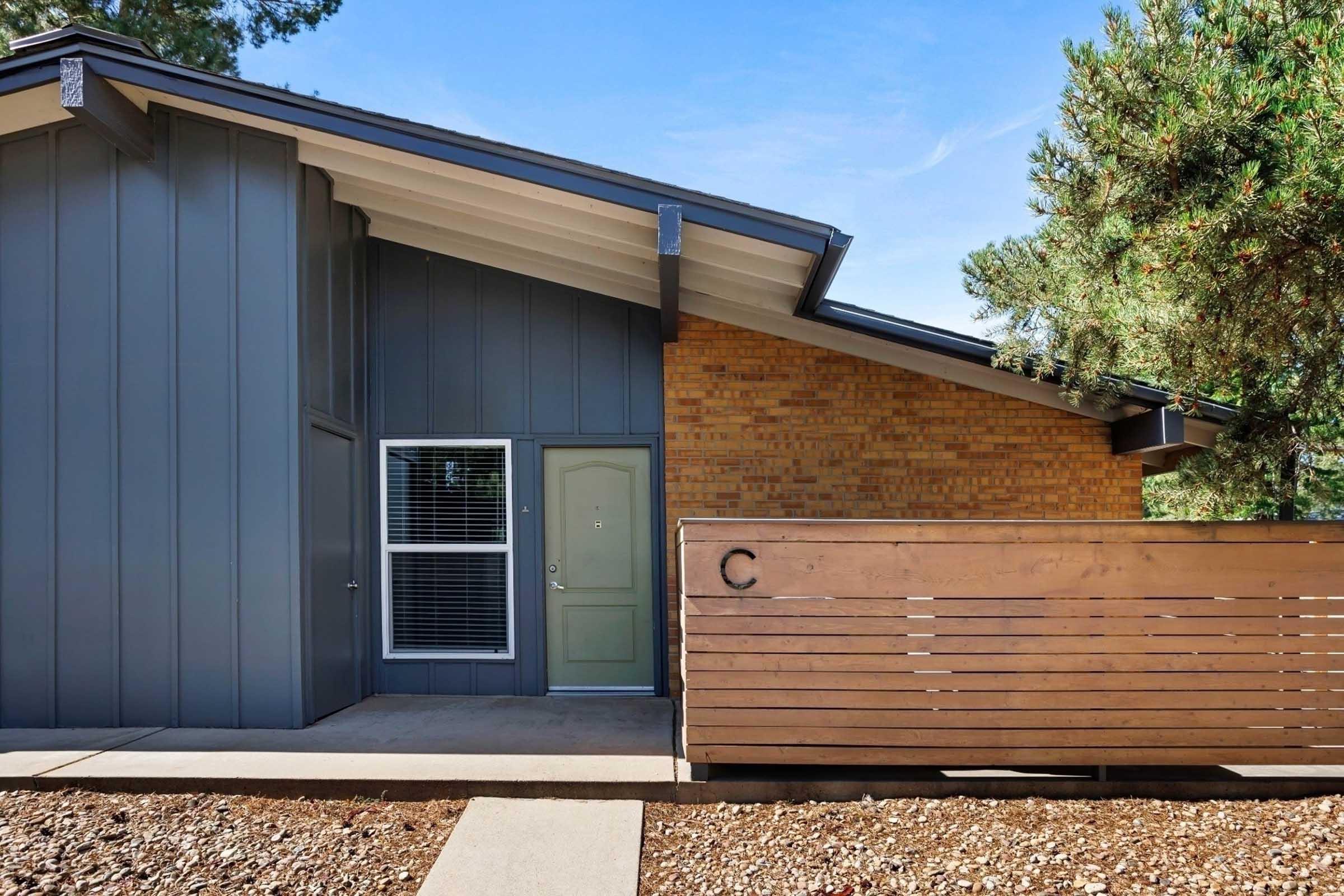 A modern, single-story house featuring a combination of gray and brick exterior. The entrance has a large wooden panel with the letter "C" displayed. There are landscaped details and a clear blue sky in the background.