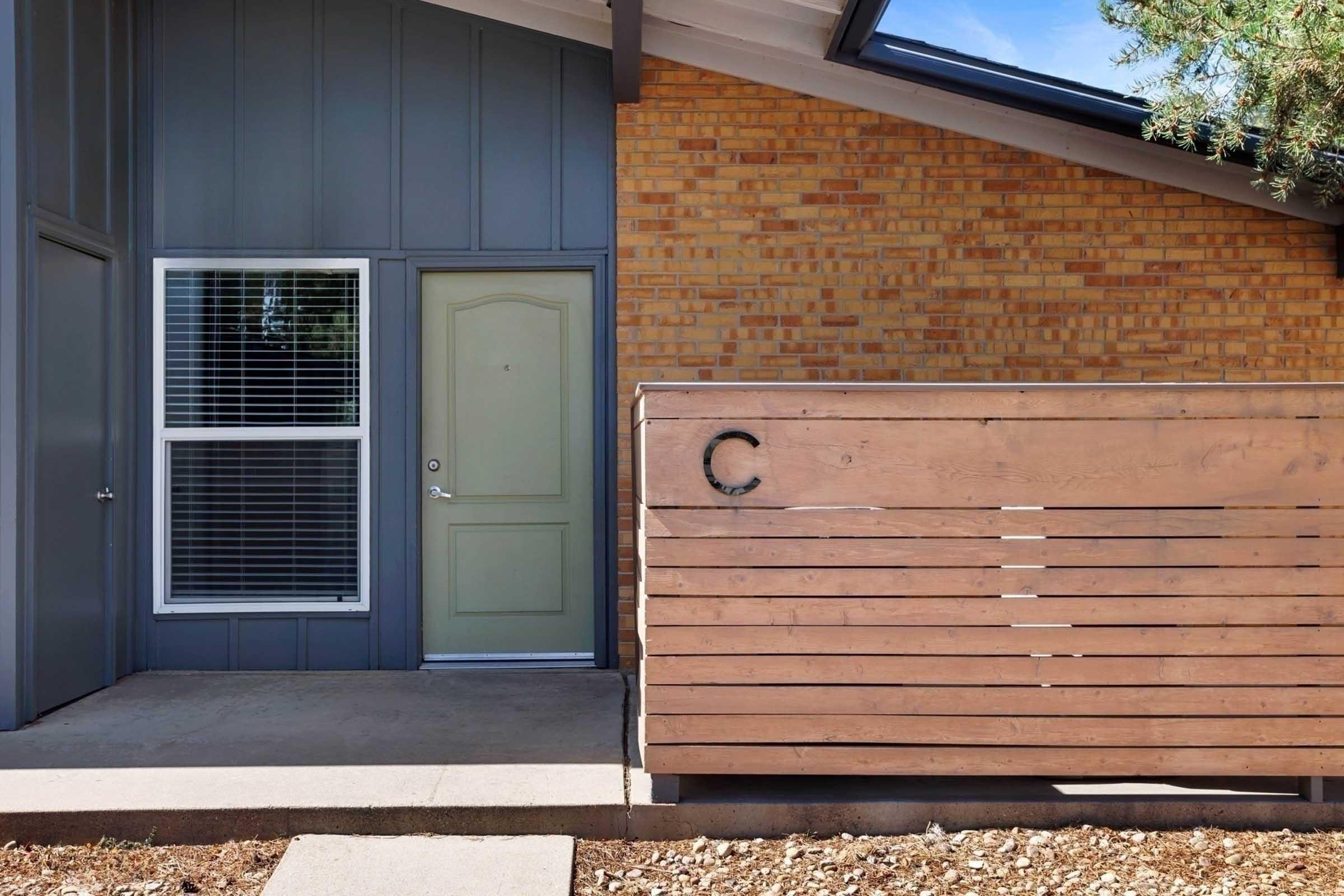 Entrance of a modern house featuring a light green door, large windows with horizontal blinds, and a wood slatted fence marked with the letter "C." The facade combines brick and contemporary lines, with a clear blue sky above and minimal landscaping around the entrance.