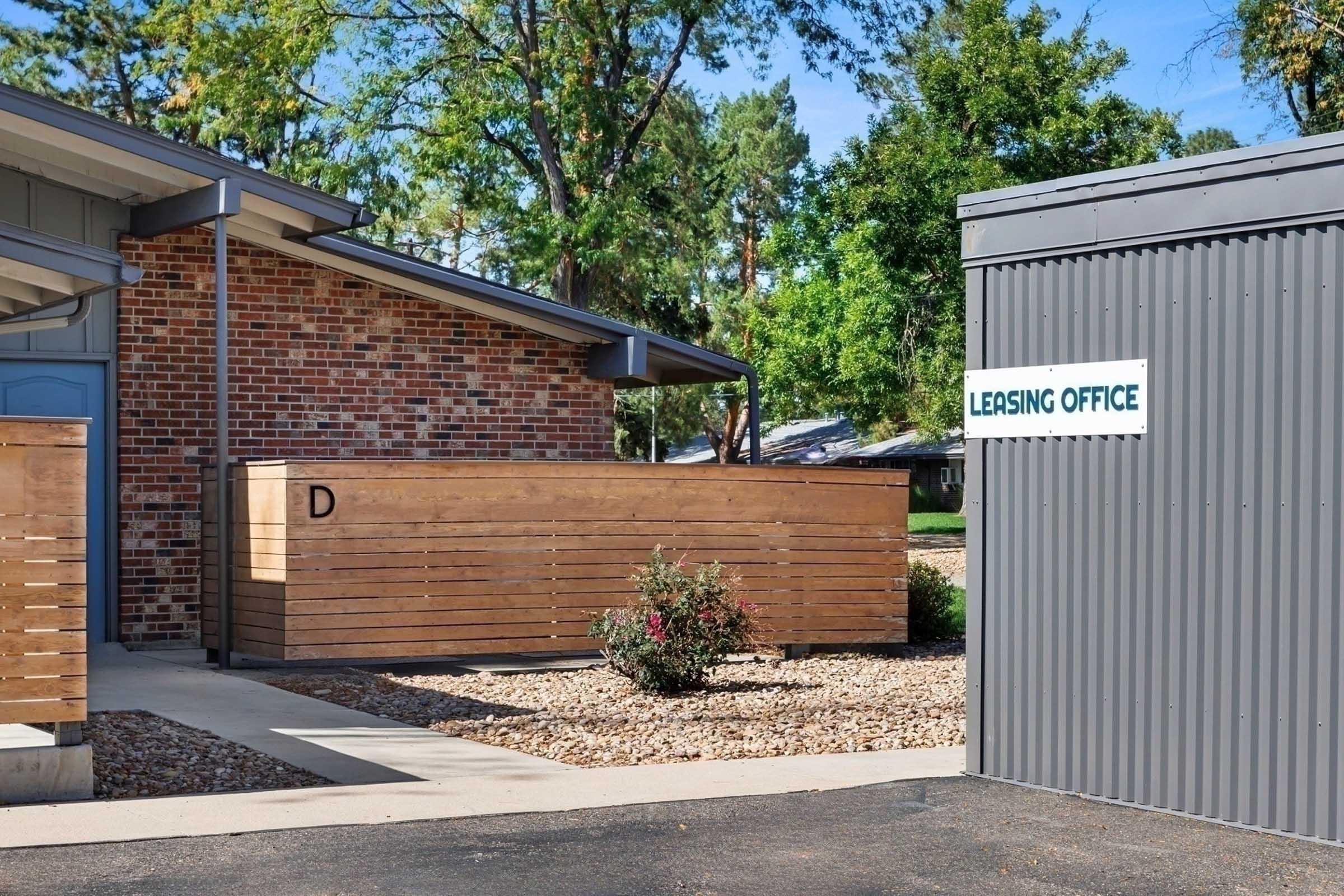 A modern leasing office building with a brick exterior and wooden accents. A sign reading "LEASING OFFICE" is visible on a gray structure nearby. The area is landscaped with small bushes and trees in the background, suggesting a pleasant environment for potential renters.