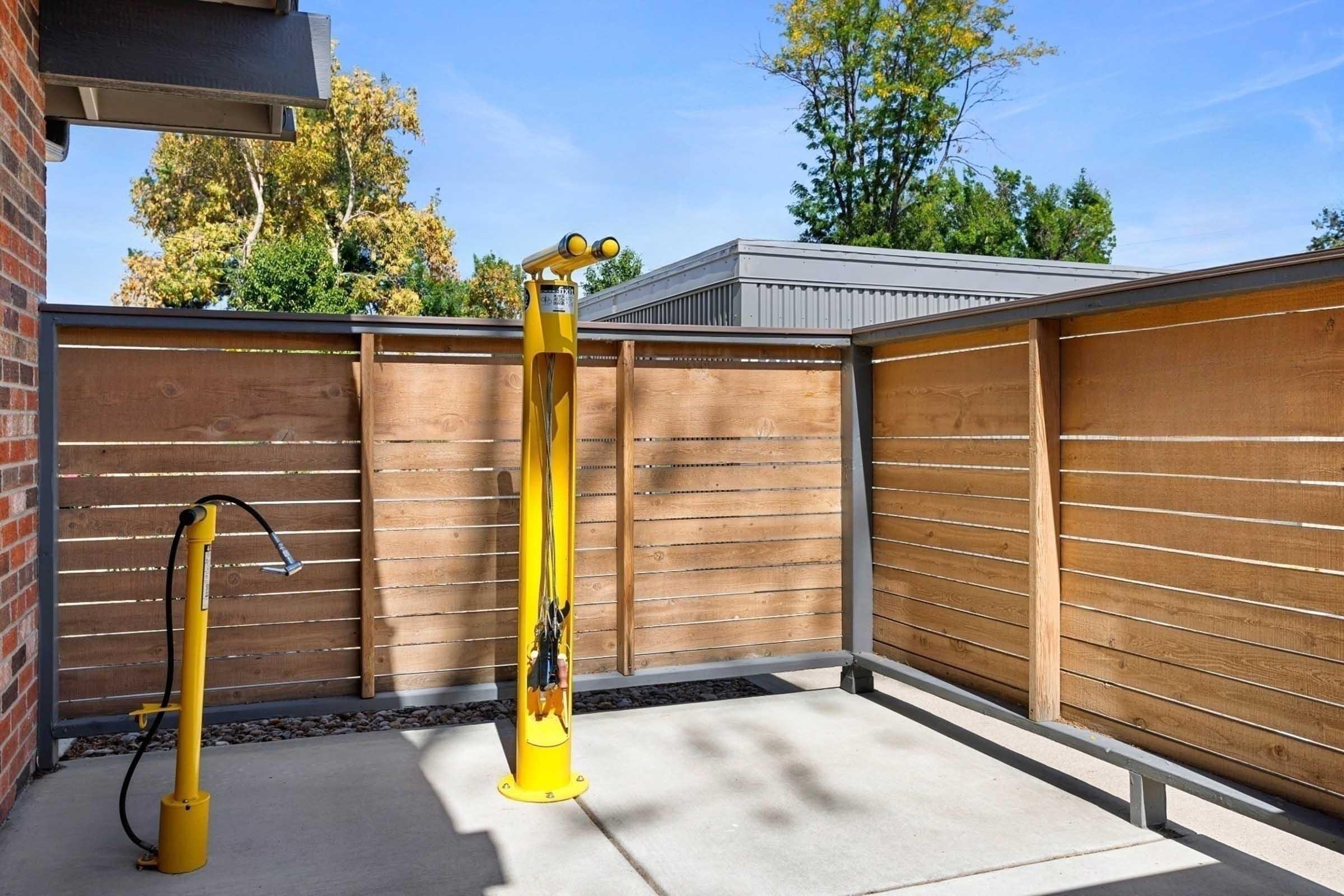 A small outdoor area featuring two cylindrical yellow posts. One post has a hose attached, possibly for water, while the other appears to be a vacuum or air station. The space is enclosed by wooden slats and surrounded by greenery in the background, under a clear blue sky.