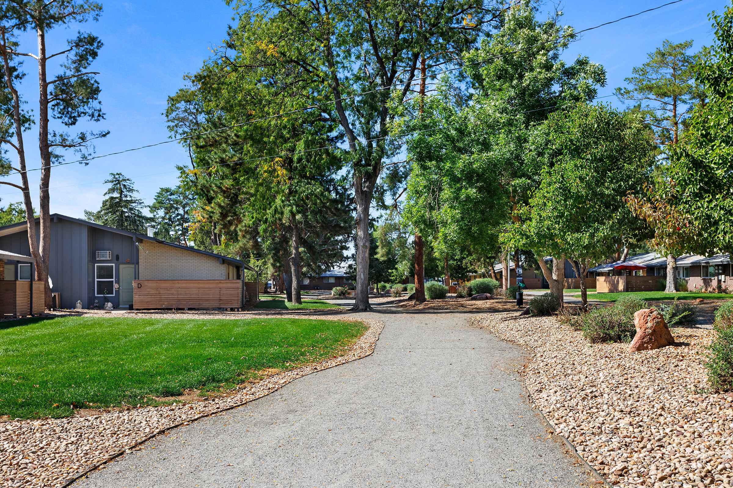 A quiet pathway winding through a landscaped area with green grass, trees, and gravel. On one side, a building with a wooden facade is visible, while houses can be seen in the distance. The scene suggests a peaceful neighborhood or park setting, ideal for walking and enjoying nature.