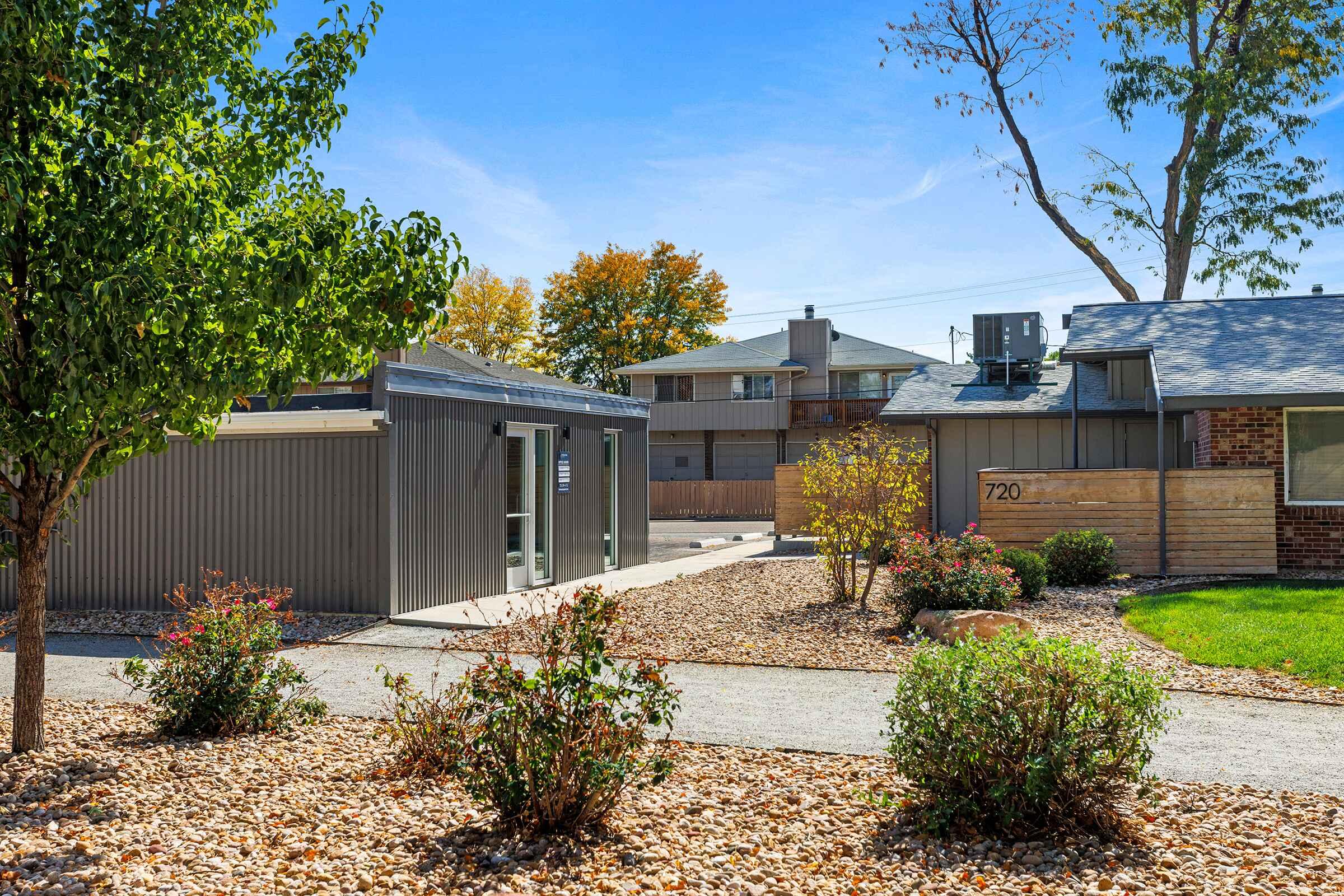 A modern building with a gray exterior and large windows sits beside a pathway. Colorful shrubs and decorative stones line the path, leading to a nearby residential area with multiple buildings. Trees with autumn foliage add a vibrant touch to the scene under a clear blue sky.