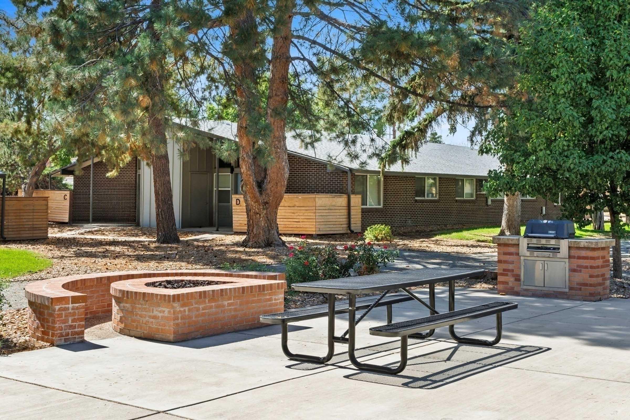 A landscaped outdoor area featuring a picnic table on a concrete slab, surrounded by trees and bushes. In the background, there is a brick building with several windows and wooden planters. To the right, a stone fire pit and a barbecue grill are visible, creating a social gathering space.