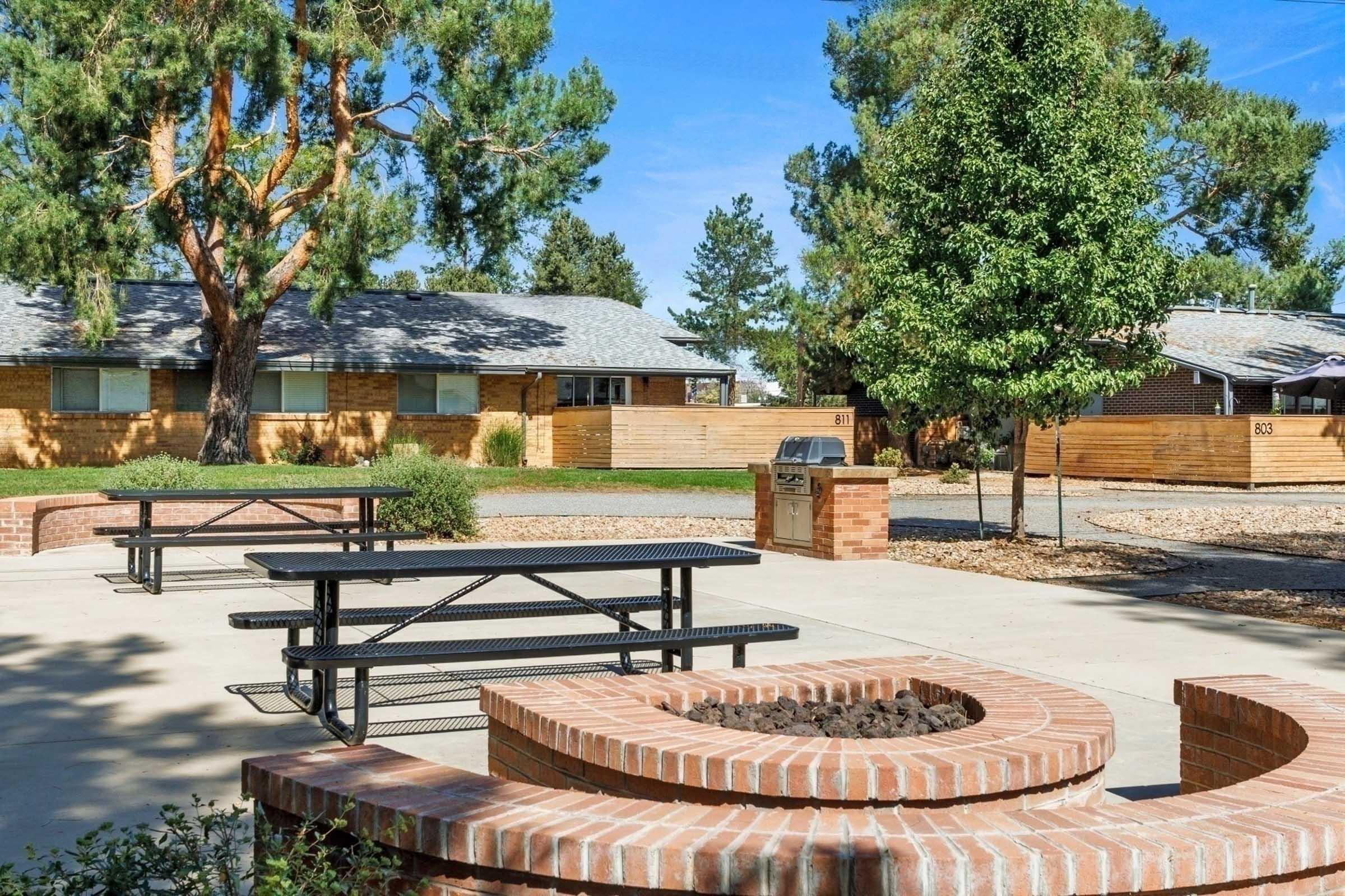 A landscaped outdoor area featuring picnic tables, a brick fire pit, and a grill. Surrounding trees provide shade, and nearby buildings are partially visible behind wooden fences, all set against a clear blue sky. The space is ideal for gatherings or relaxation.