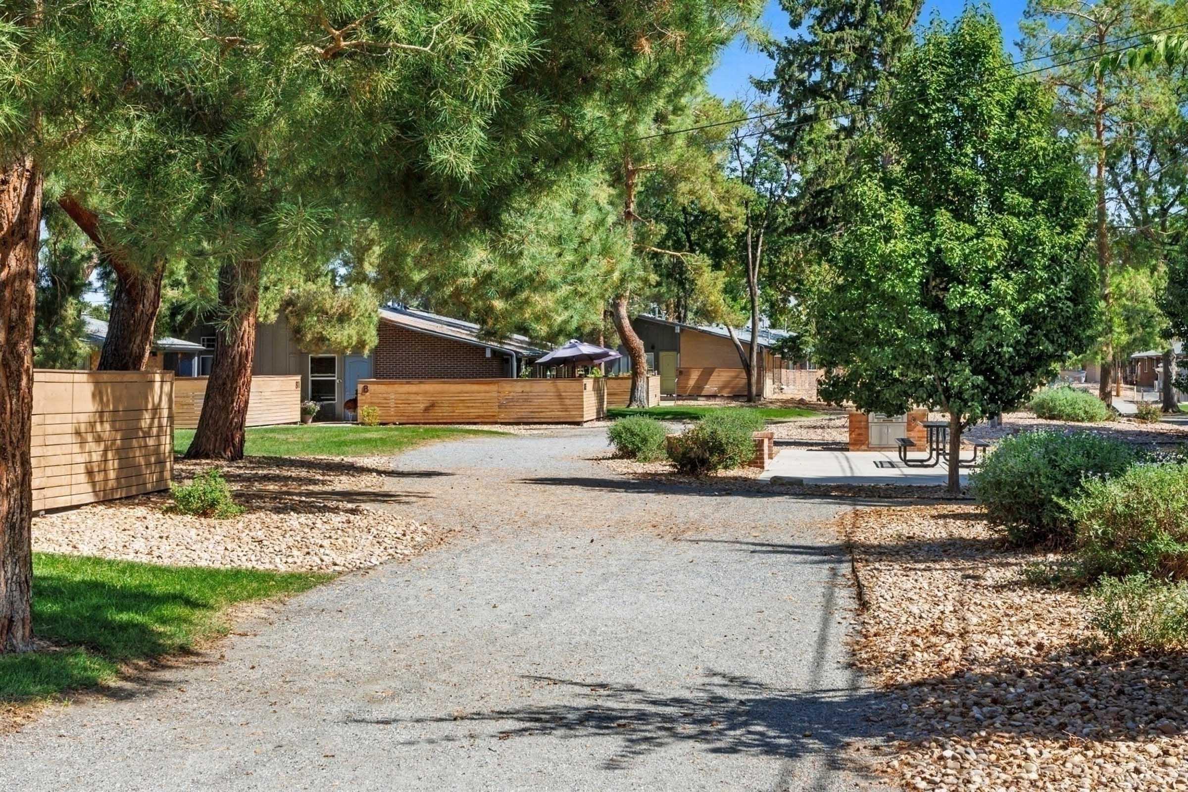 A peaceful pathway curves through a landscaped area lined with trees and shrubs. On either side, wooden fences create a cozy atmosphere, with buildings visible in the background. Sunlight filters through the trees, enhancing the tranquil setting, making it an inviting space for leisurely strolls.