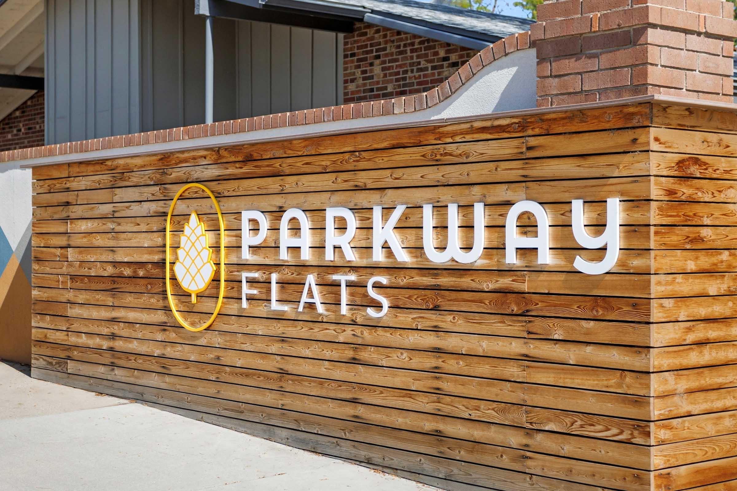 Wooden sign for Parkway Flats featuring the name in large white letters along with a stylized pinecone logo, set against a rustic wood backdrop. The sign is prominently displayed in an outdoor setting, contributing to an inviting atmosphere.