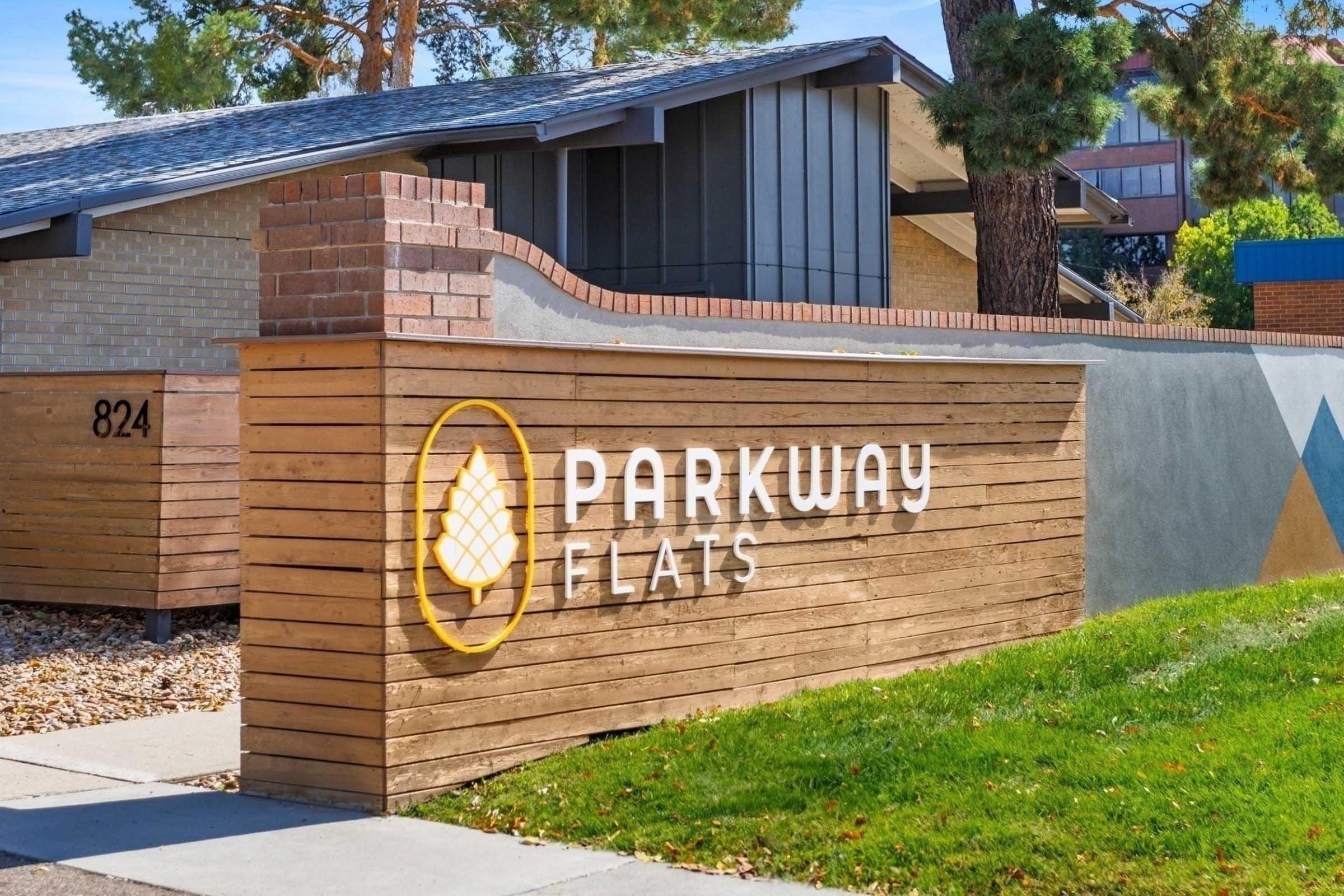Sign for Parkway Flats featuring a modern design with a wooden texture. The sign showcases the name "PARKWAY" in large, bold letters above the word "FLATS." The background includes a landscaped area with trees and a building facade in the background.