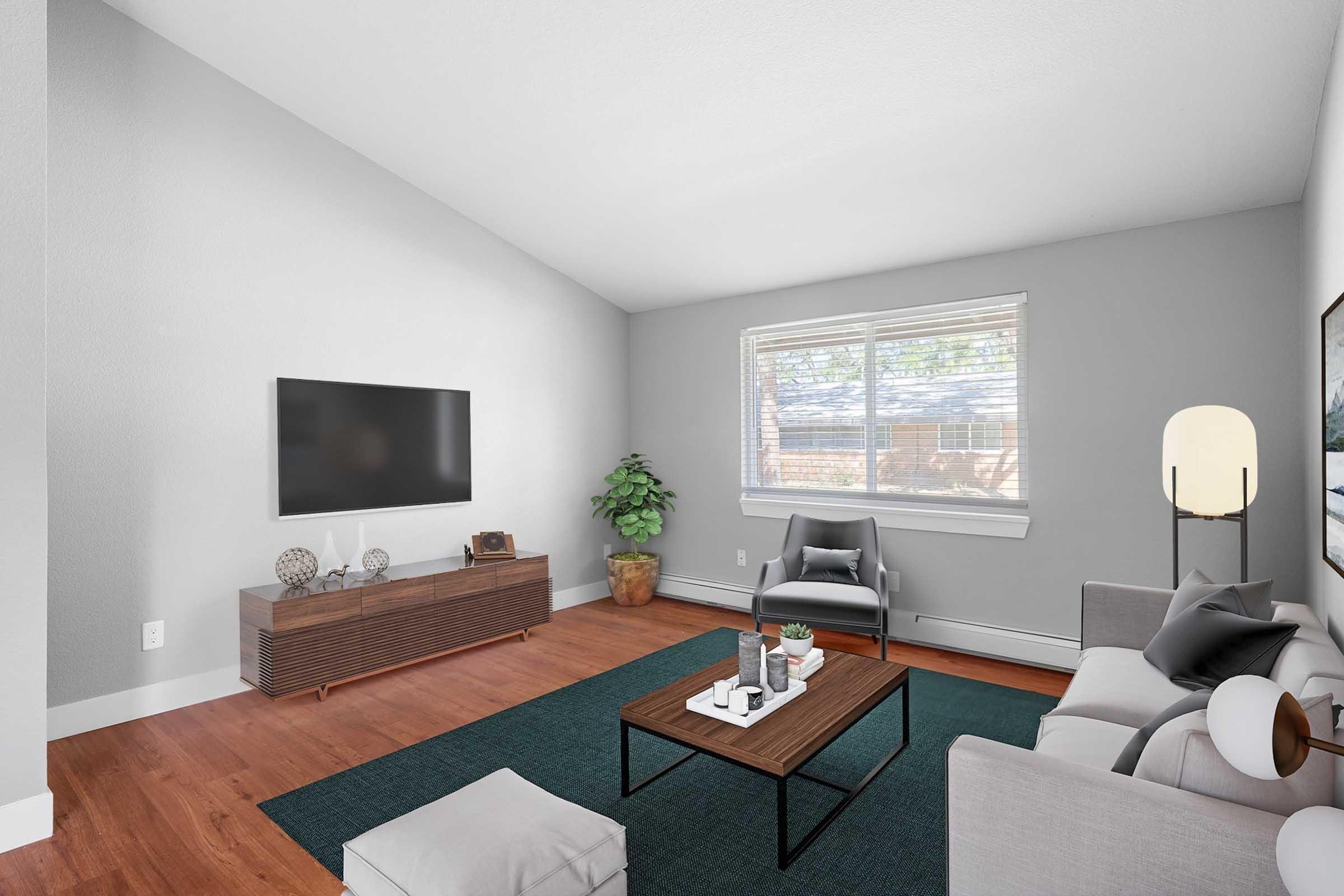 A modern living room featuring light gray walls, a flat-screen TV mounted on the wall, a stylish coffee table, and comfortable seating. A large window allows natural light to fill the space, while a potted plant adds a touch of greenery. The flooring is wooden, and a dark green area rug complements the decor.