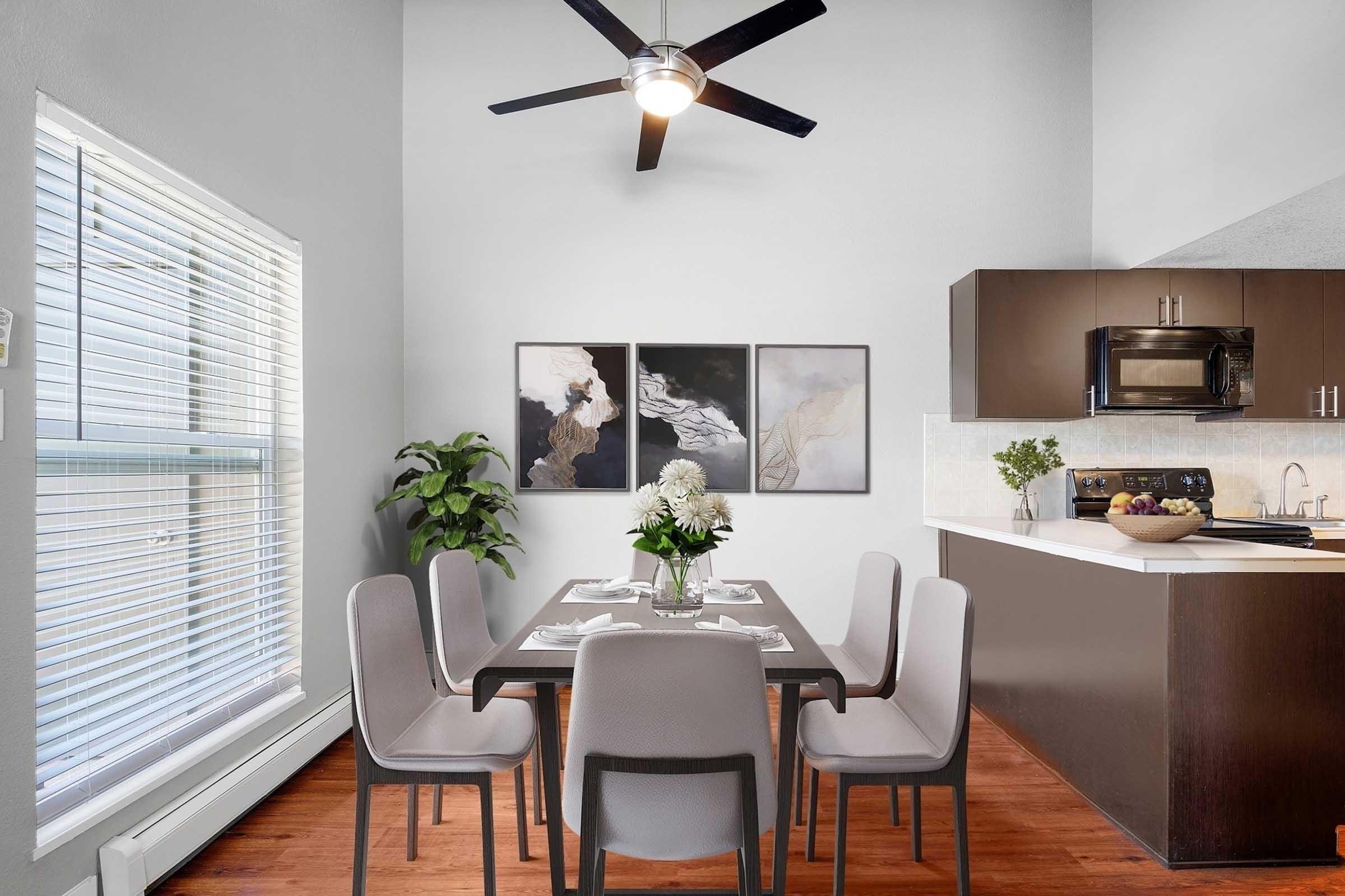 A modern dining area featuring a dark wood table set for six, surrounded by gray upholstered chairs. A ceiling fan is above, and there are two abstract art pieces on the wall. A potted plant sits in the corner, and a kitchen area is visible in the background with dark cabinets and a stainless steel appliance.