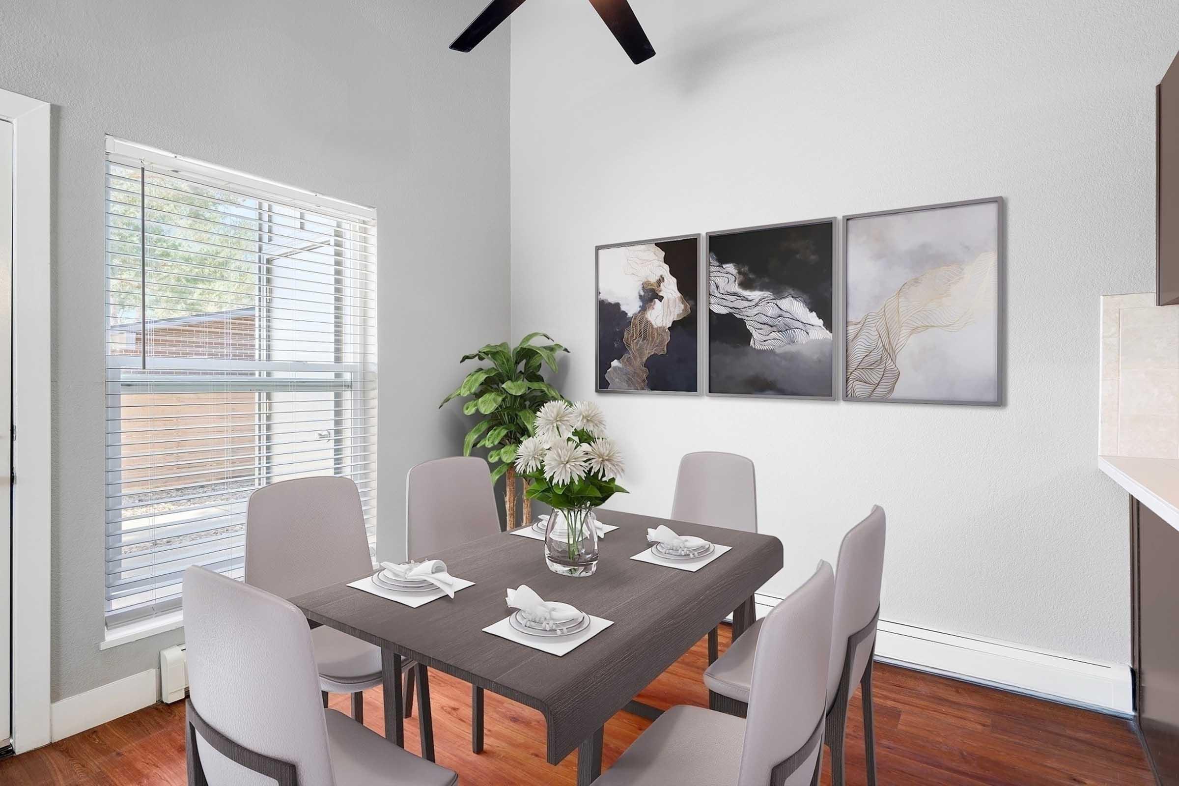 A modern dining area featuring a gray table with six chairs, set with white plates and napkins. A vase of white flowers is the centerpiece. The walls are painted light gray, and there are three abstract art pieces on the wall. A window lets in natural light, with blinds partially open.