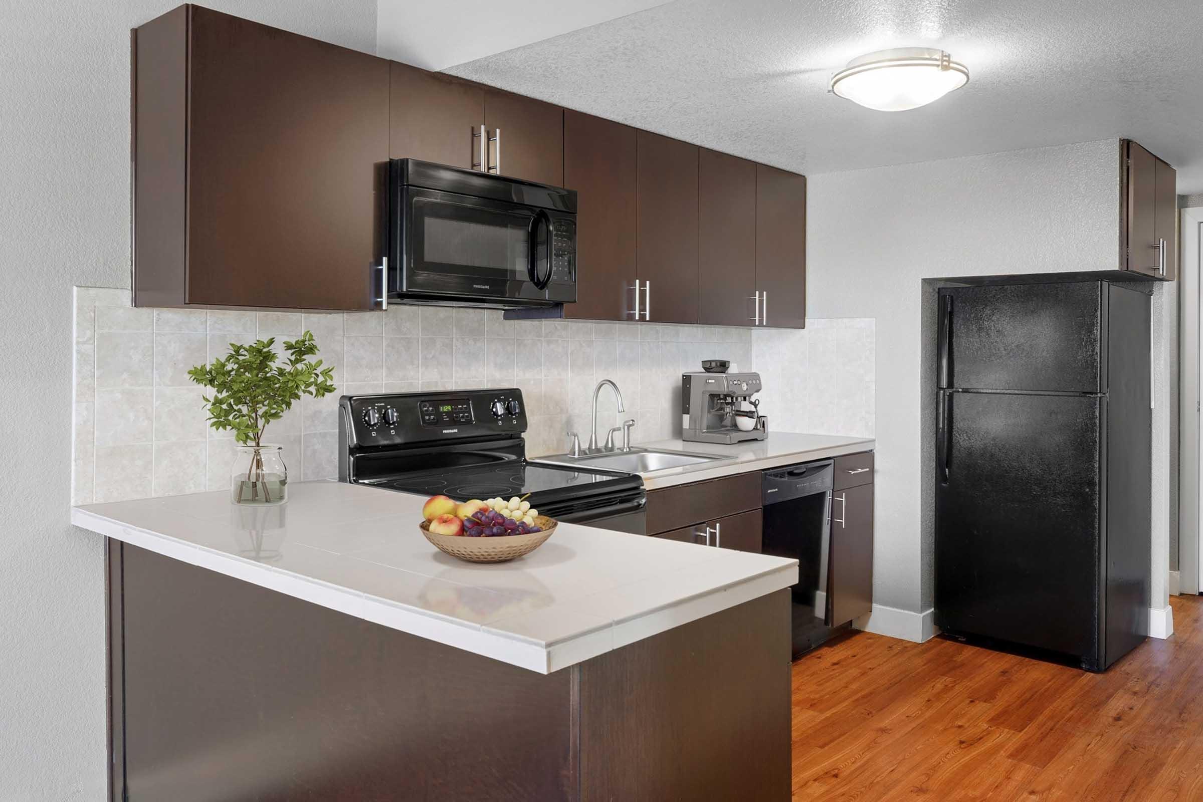 Modern kitchen featuring dark cabinets, a black microwave, and an electric stove. A sleek countertop with a fruit bowl is in the foreground, while a black refrigerator is positioned nearby. The walls are light-toned, and there's a small sink with a faucet. Wood flooring completes the space.
