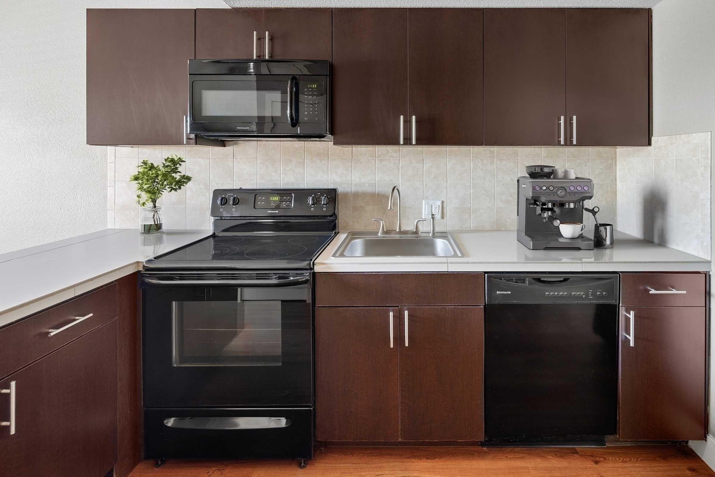 Modern kitchen featuring dark wood cabinets, a black microwave, and an electric stove. A stainless steel sink is positioned under the window, alongside a coffee machine on the countertop. A black dishwasher is also visible beneath the counter, with light-colored tiles on the walls.