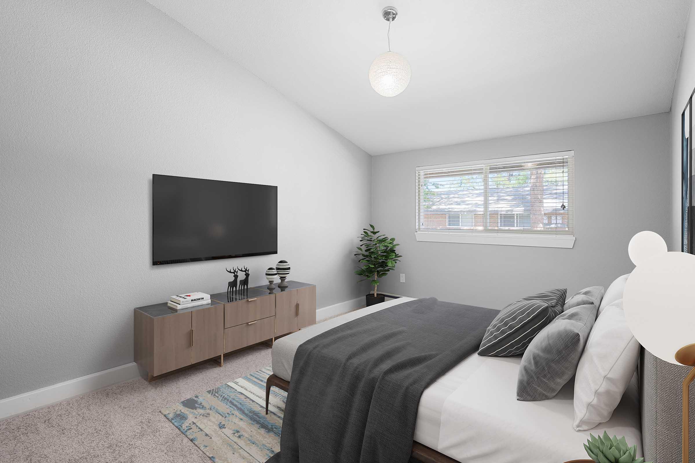 A modern bedroom featuring a bed with gray and white bedding, a wooden nightstand, and a flat-screen TV mounted on the wall. A small plant is placed near the window, and a light fixture hangs from the ceiling. The room has neutral gray walls and a cozy, inviting atmosphere.