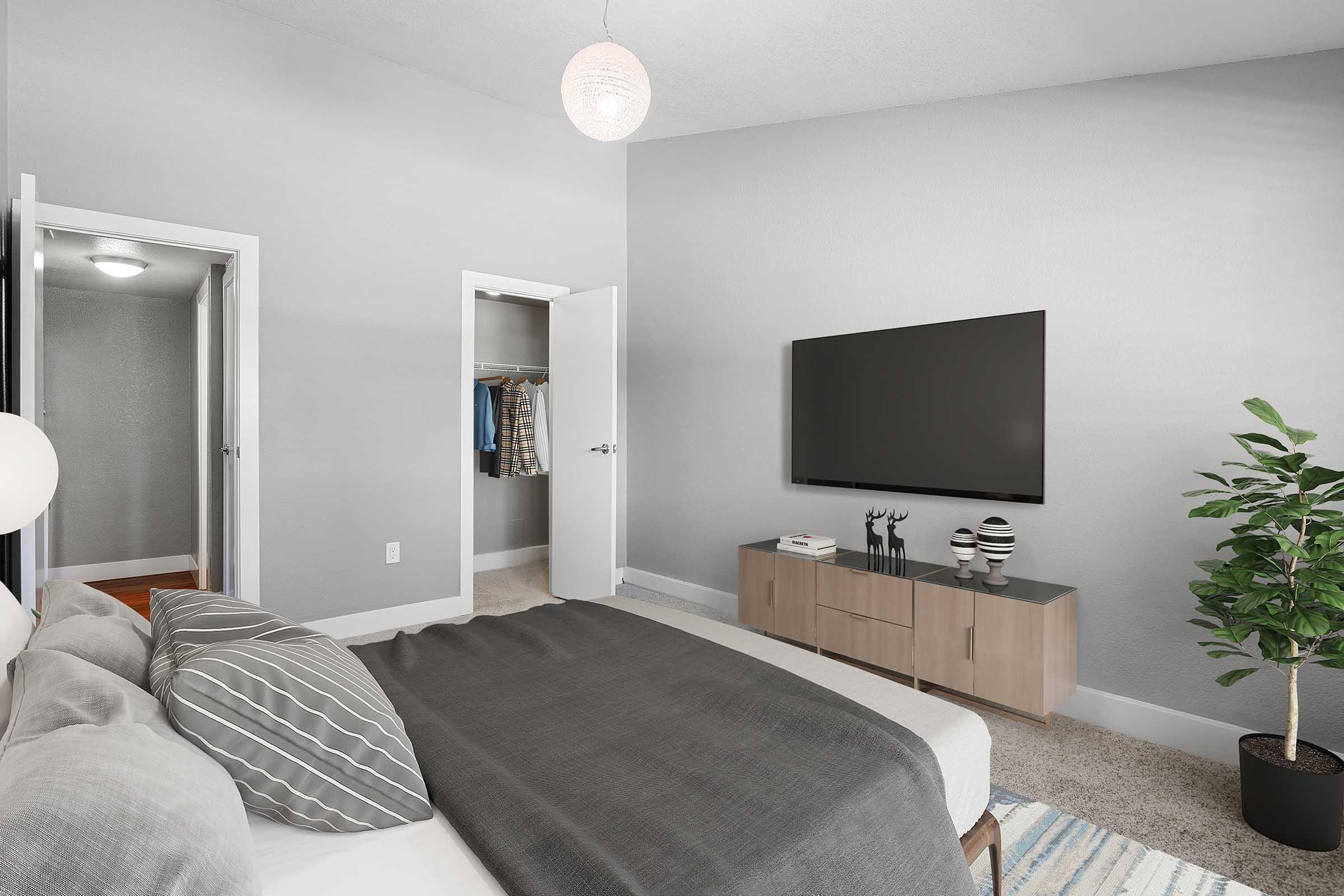 A modern bedroom featuring a gray color scheme. The focal point is a neatly made bed with decorative pillows. There's a sleek TV mounted on the wall opposite the bed, and a wooden cabinet below it. A tall, leafy plant adds a touch of greenery, while a closet door and an additional door lead to other spaces. Soft lighting creates a cozy atmosphere.