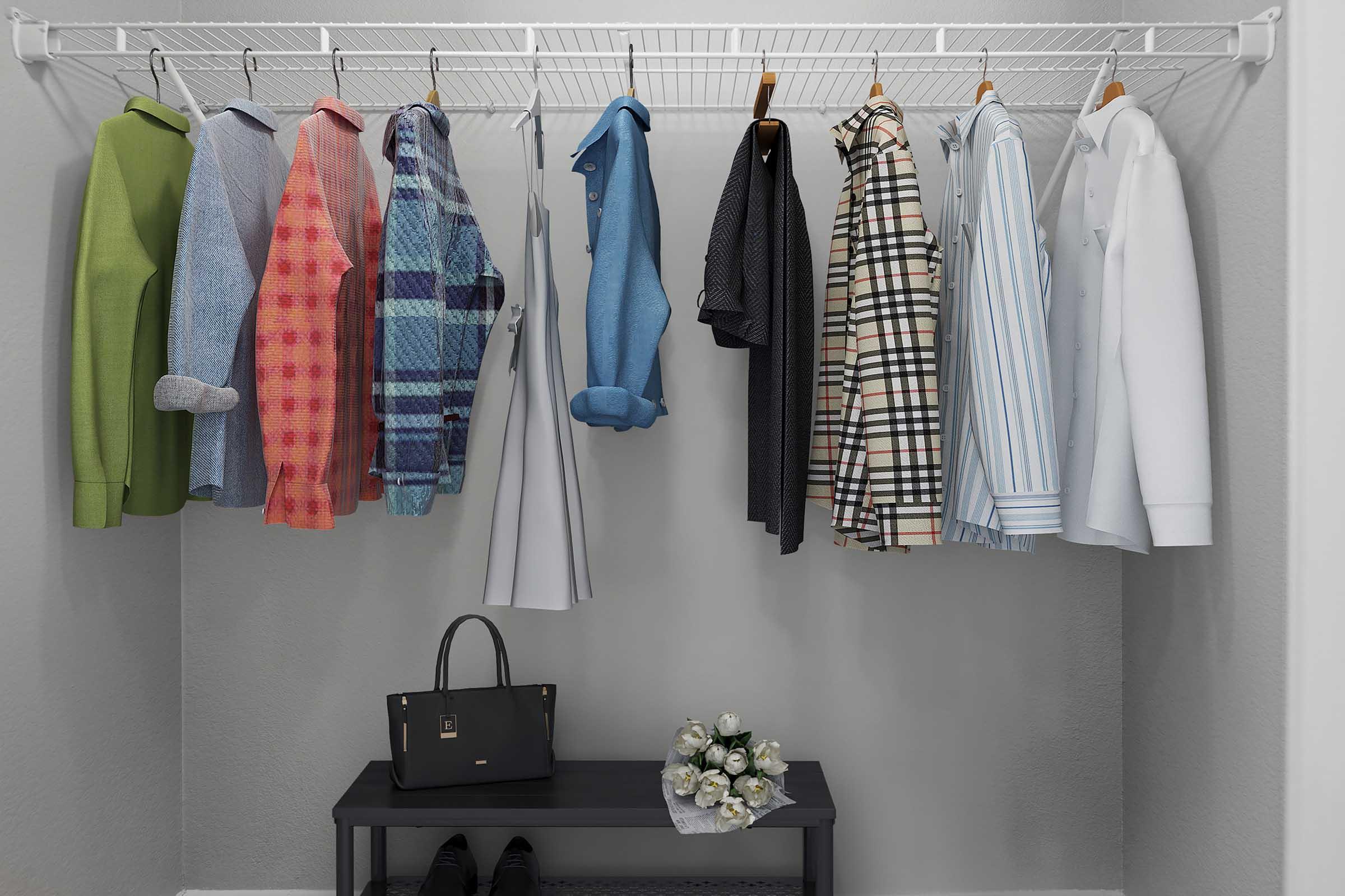 A neatly organized closet featuring a variety of shirts in different colors and patterns hanging on a rod. There is also a long skirt, a black handbag on a small bench, and a vase with flowers positioned on the bench, creating a tidy and stylish space.