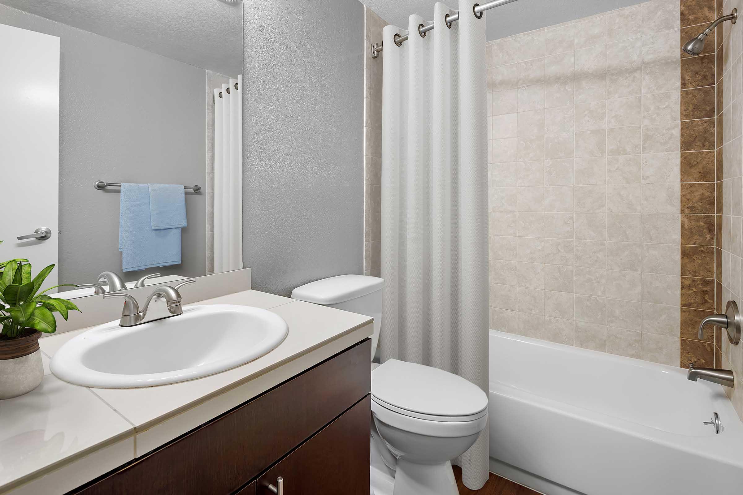A clean and modern bathroom featuring a white sink with a mirror above, a bathtub with beige tiles, a shower curtain, and a small potted plant. Light gray walls complement the overall design, with a neatly folded blue towel hanging nearby.