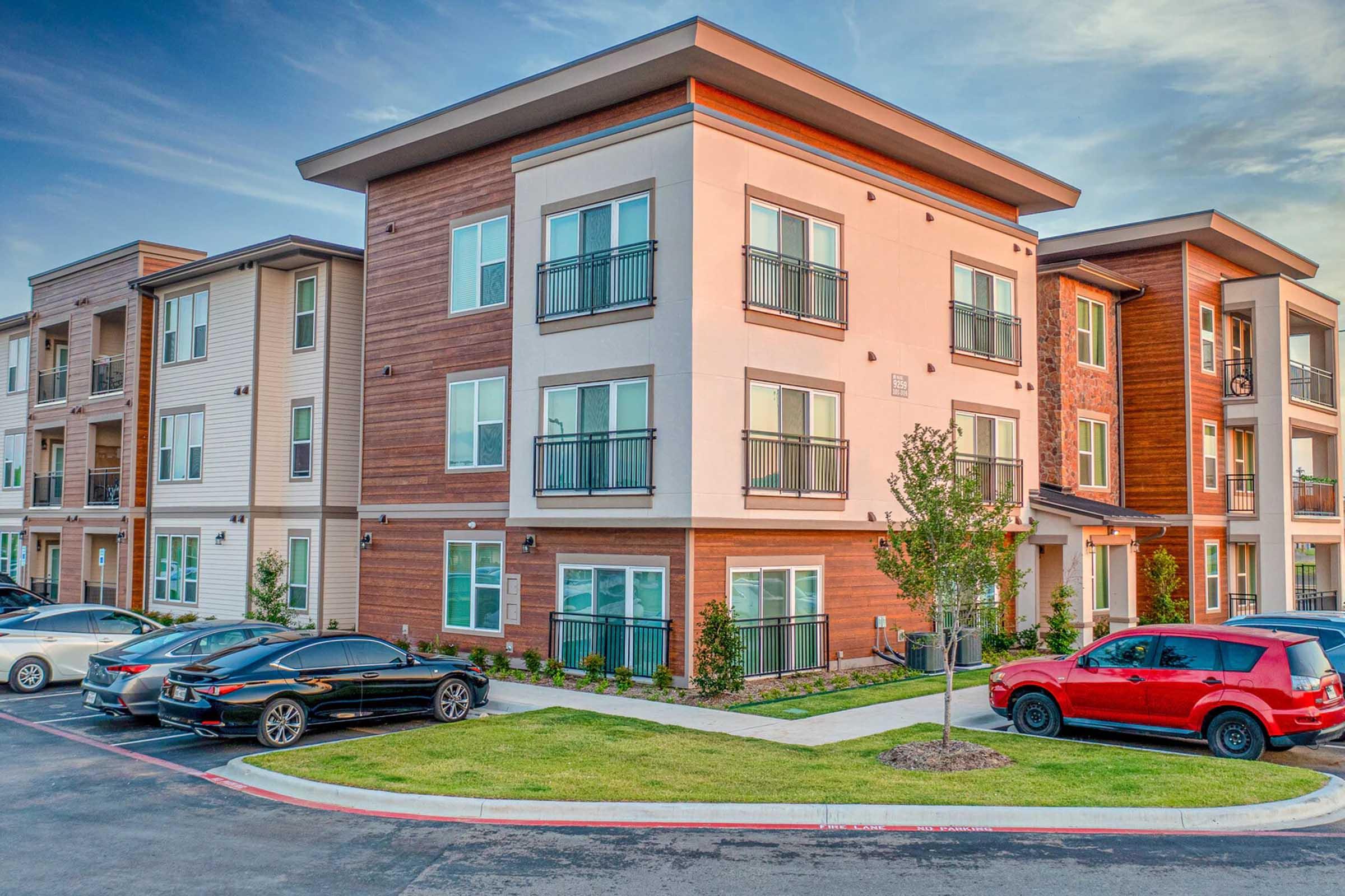 Modern apartment complex featuring a three-story building with a mix of light and dark wood paneling. The facade includes balconies, large windows, and landscaped green space. Several cars are parked in front, with a spacious driveway and a clear sky in the background.