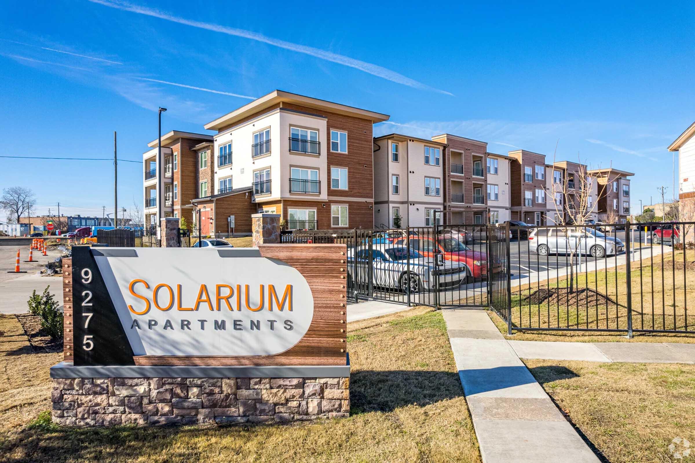 Sign for Solarium Apartments with the address 9275, set in front of multi-story residential buildings. The complex features modern architecture with balconies, surrounding parking, and landscaped grounds under a clear blue sky.