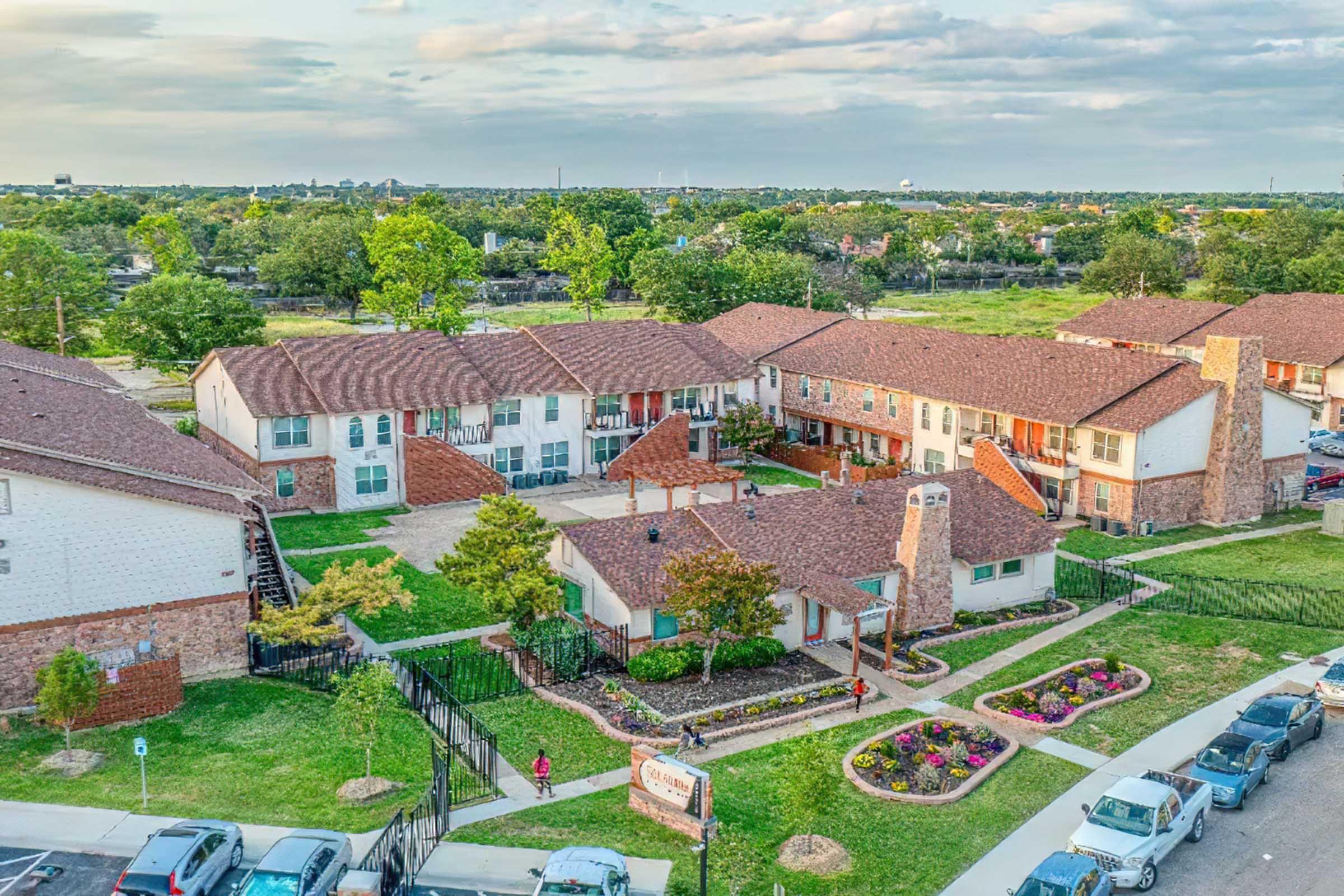 Aerial view of a residential complex showcasing multiple buildings with red roofs, well-maintained grassy areas, and landscaping. The scene includes pathways, trees, and flower beds, set against a backdrop of green fields and a cloudy sky.